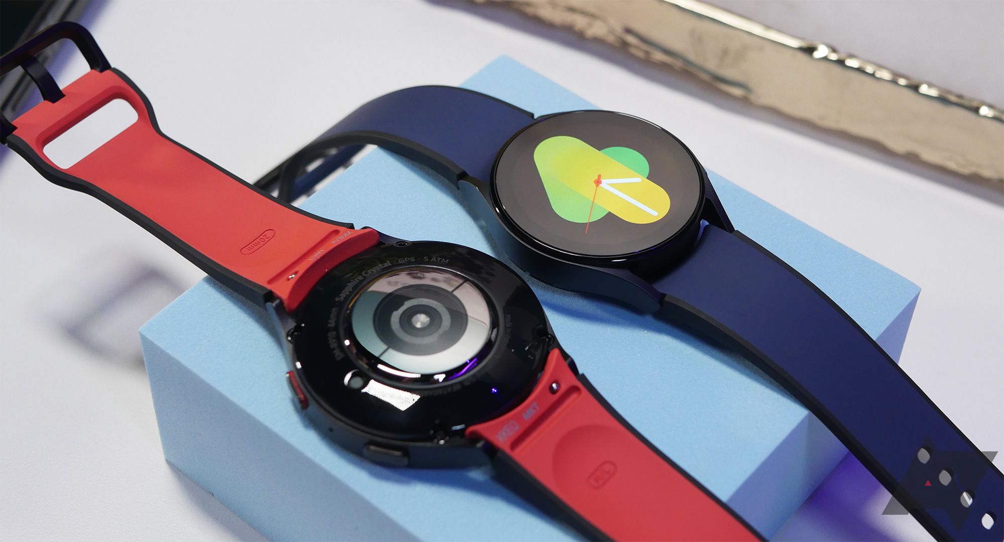 dois smartwatches estão em um bloco azul pastel.  Um deles possui display iluminado;  o outro está de bruços. 