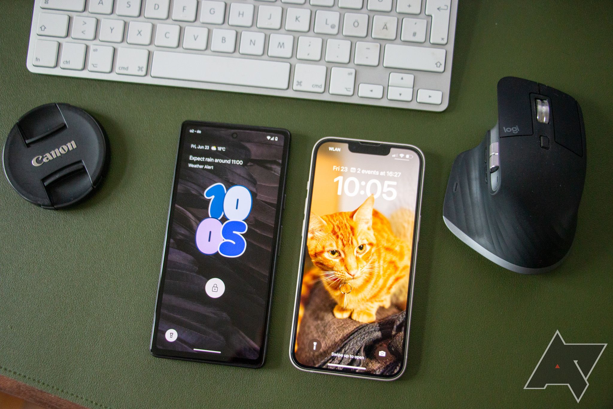 Um Google Pixel 7a e um iPhone 13 estão lado a lado em uma superfície de couro verde, com uma tampa de câmera Canon, um teclado Apple Magic e um mouse Logitech artisticamente dispostos ao redor deles