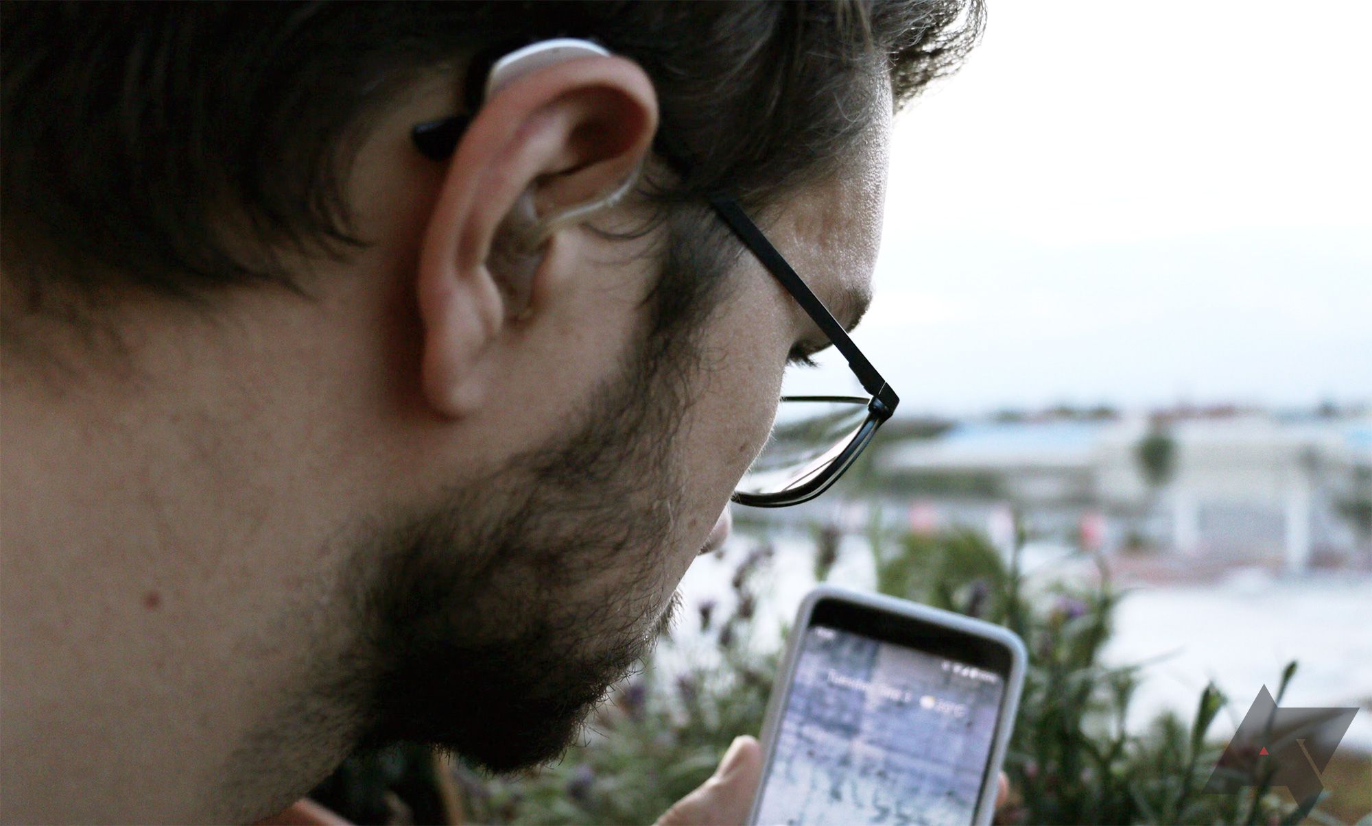 Um homem está de costas para a câmera, com um aparelho auditivo visível no ouvido.  Ele está olhando para um telefone com a tela ligada.