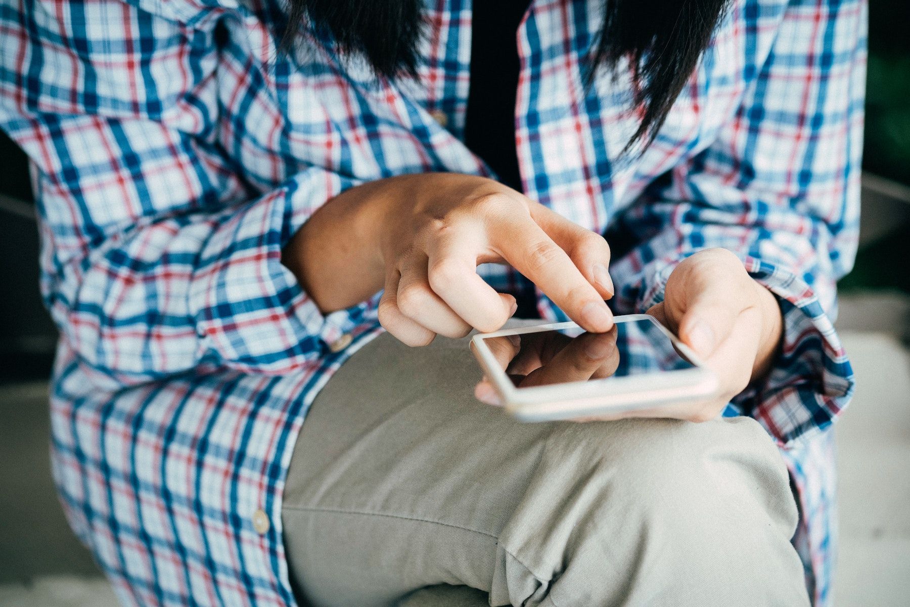 Uma pessoa vestindo uma camisa xadrez e calça cáqui segura um telefone na mão esquerda enquanto o dedo indicador direito toca na tela. 