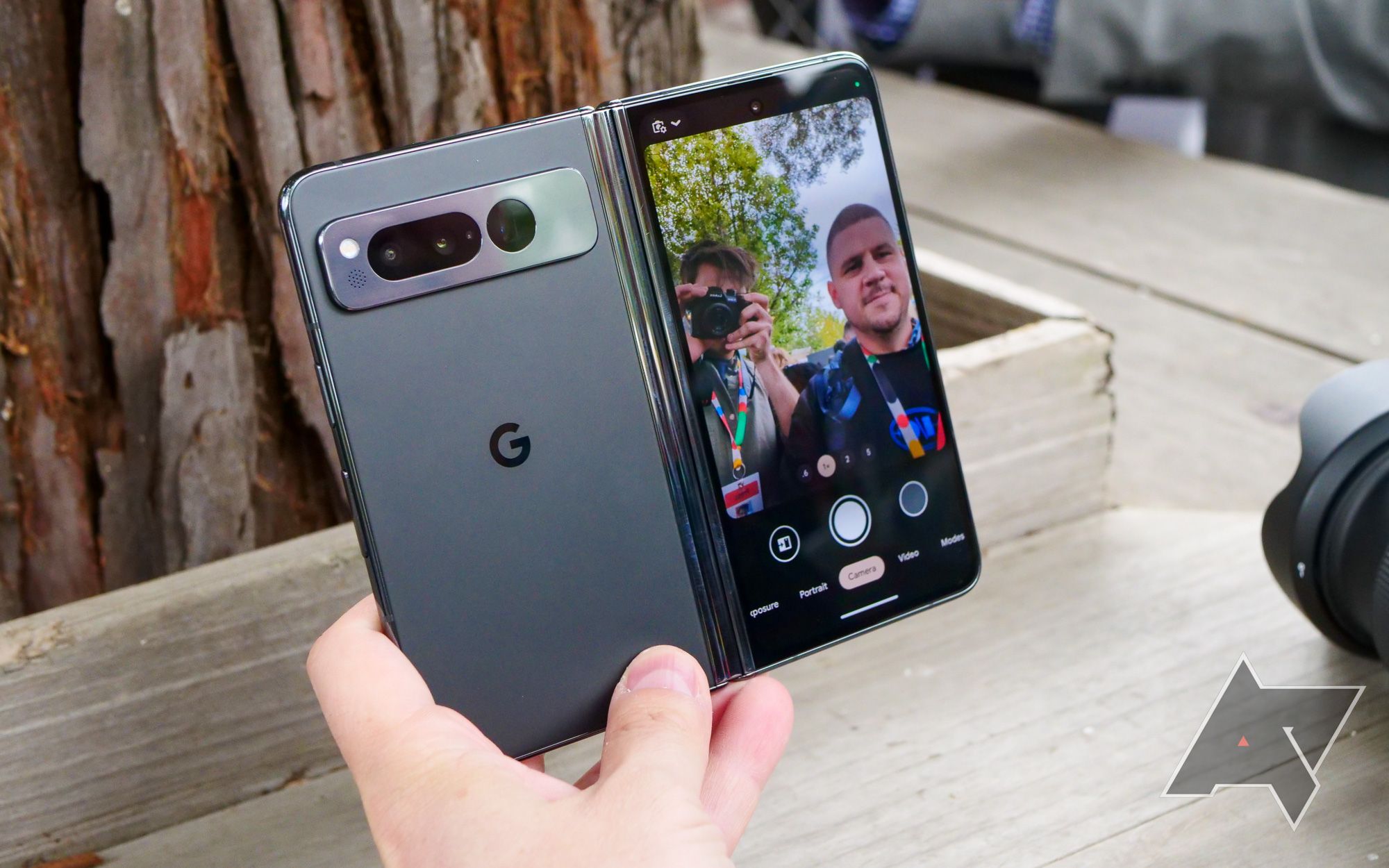 A versão preta do Google Pixel Fold é segurada com uma mão, aberta e com a parte traseira voltada para a câmera.  A tela externa é ligada, mostrando o visor da câmera e a pessoa que tira a foto.