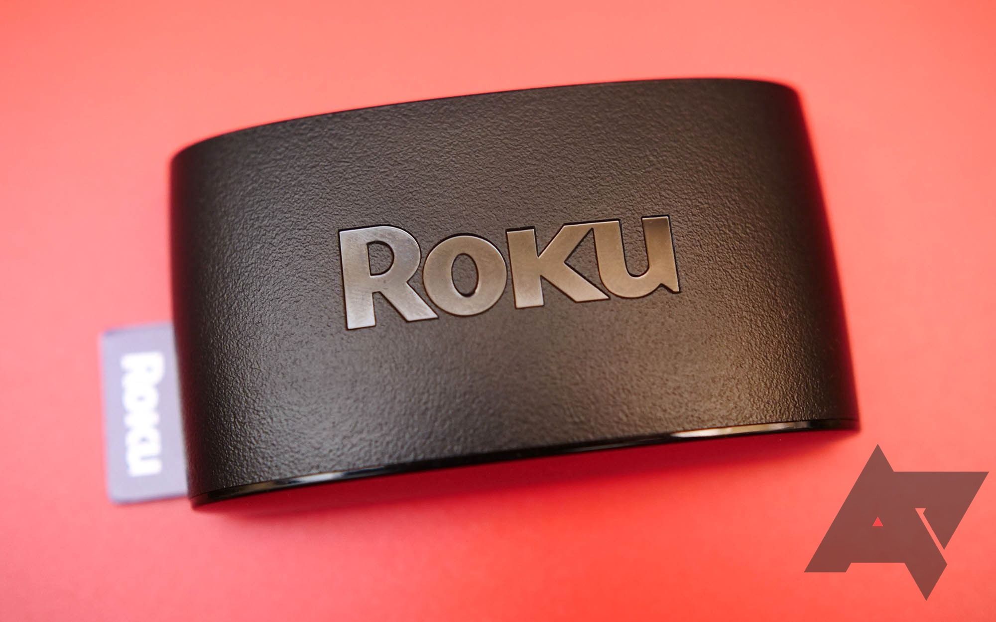 Um dispositivo Roku preto em um fundo vermelho com o logotipo do Android Police no canto inferior direito