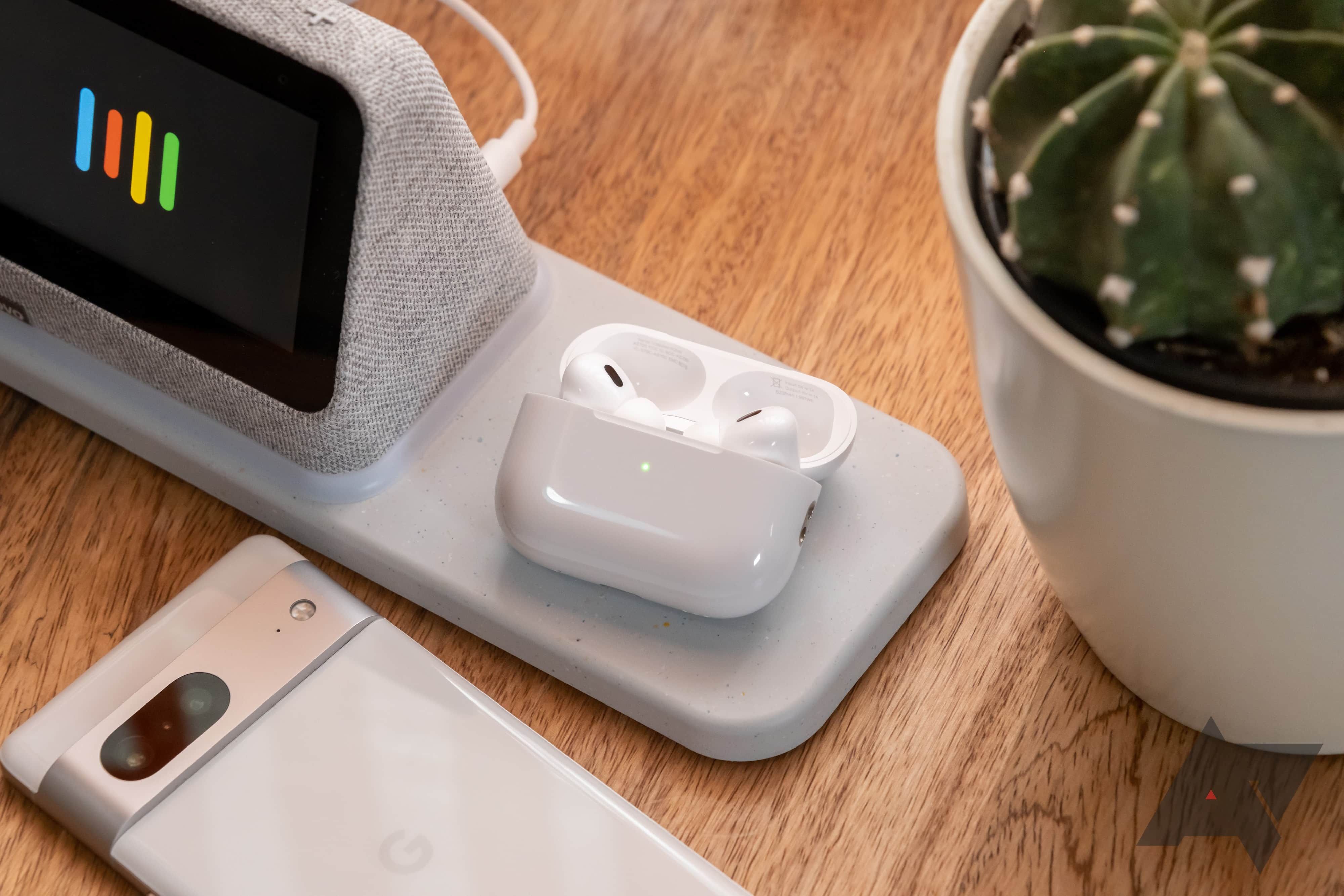 Fones de ouvido sem fio no estojo de carregamento sobre uma mesa com um smartphone, um display inteligente e um pequeno cacto.