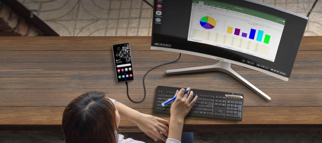 Uma pessoa sentada em frente a um monitor exibindo gráficos, um teclado e um smartphone conectado.