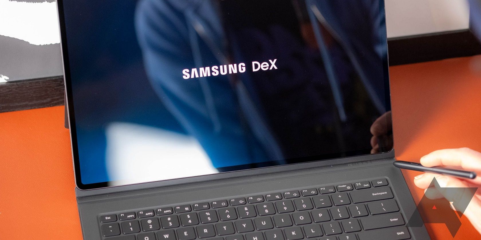 A imagem mostra um tablet Samsung com um teclado conectado, aberto, com as palavras Samsung DeX exibidas na tela. Uma mão segura uma caneta no canto inferior direito em direção à tela.