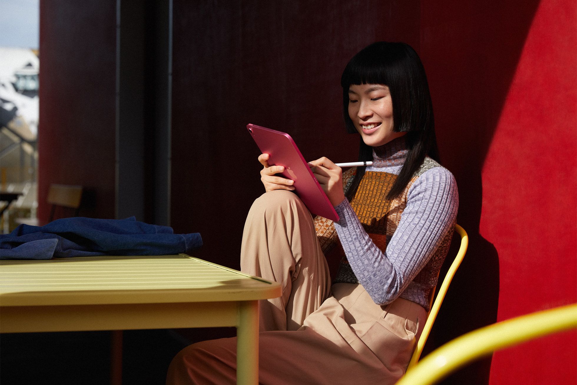 Uma pessoa está sentada em uma cadeira com fundo vermelho colorido segurando um iPad enquanto desenha nele e sorri.