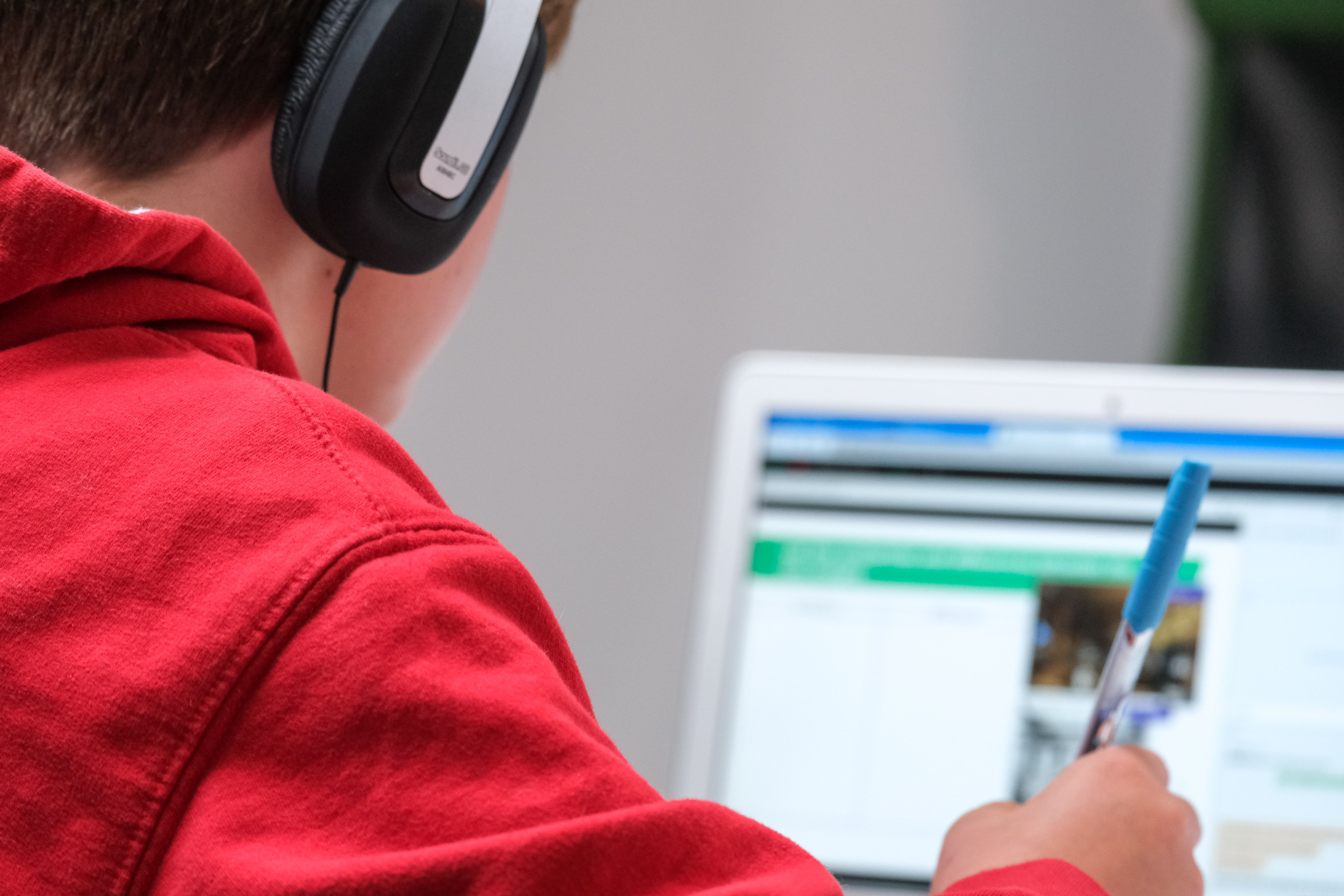 Criança vista de trás usando um moletom vermelho e fones de ouvido.  A criança tem uma caneta na mão direita e um laptop ao longe. 