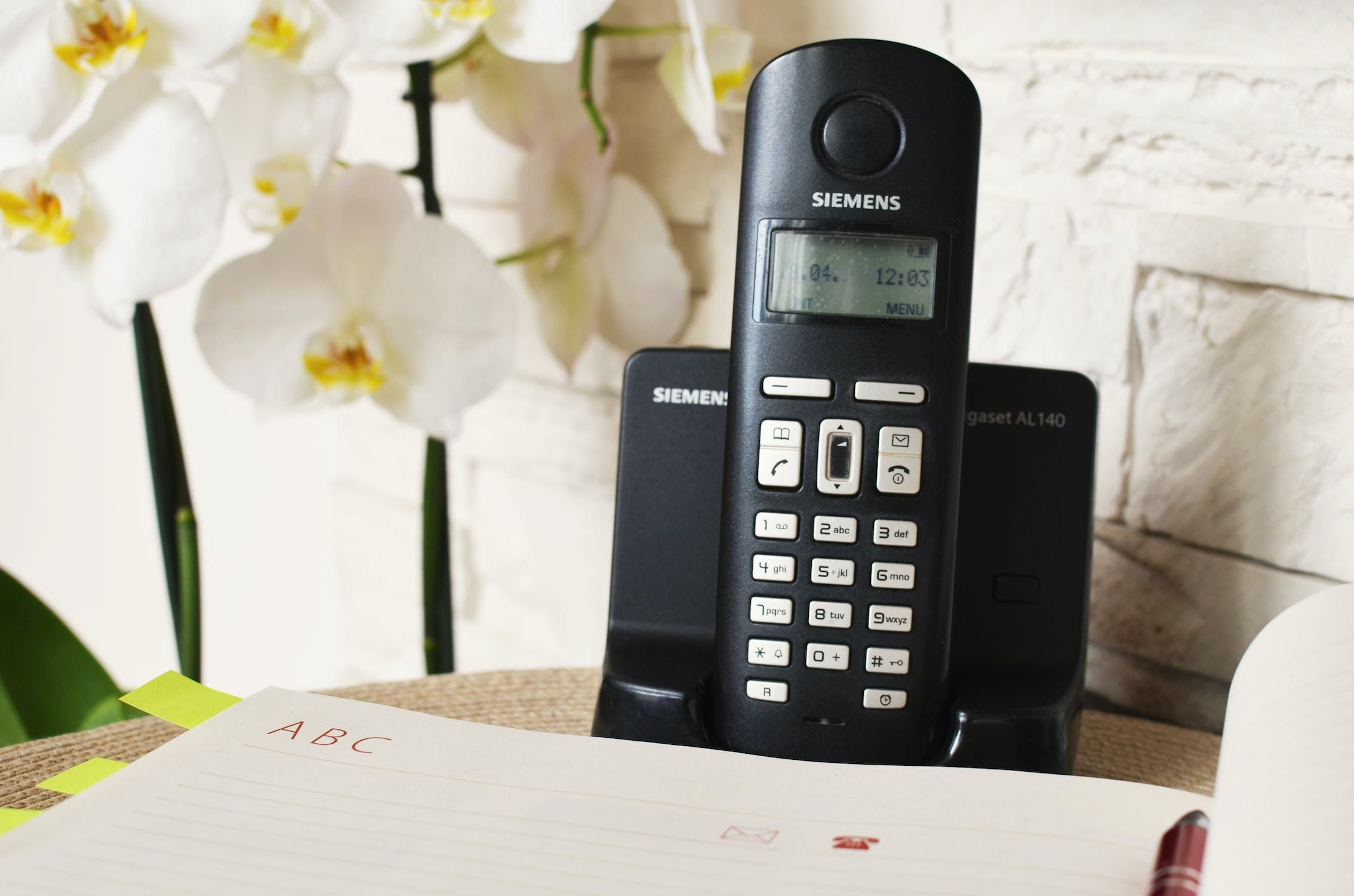 Um telefone fixo preto em uma mesa ao lado de um bloco de notas e flores brancas.