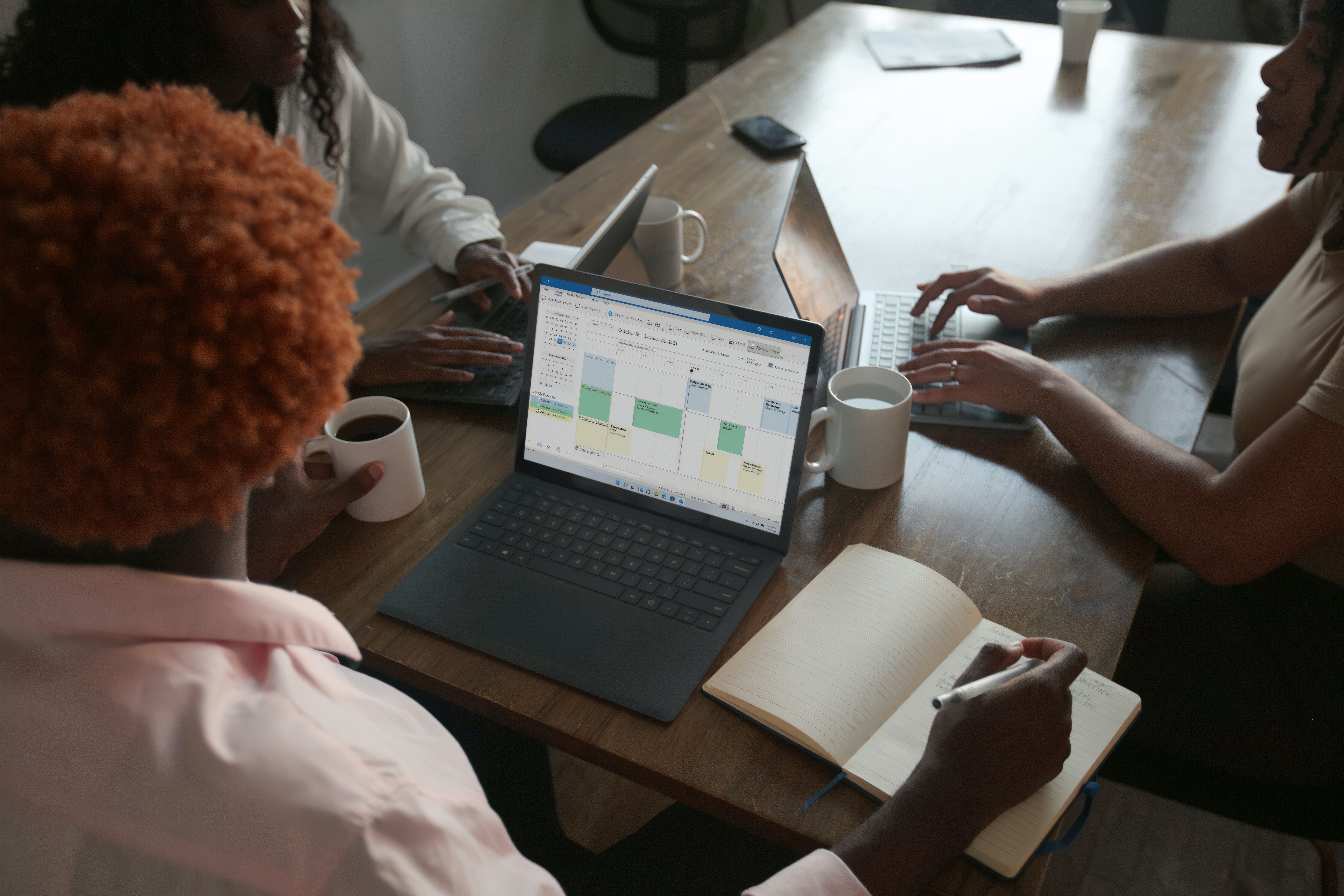 A tela de um laptop mostra uma planilha do Microsoft Excel em uma mesa cercada por funcionários. 