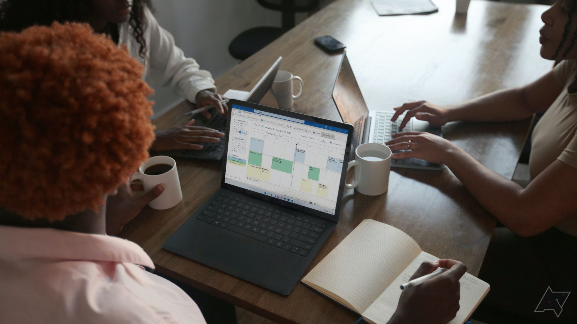Três pessoas compartilham uma mesa para usar laptops, uma delas tem o Microsoft Excel aberto na tela.