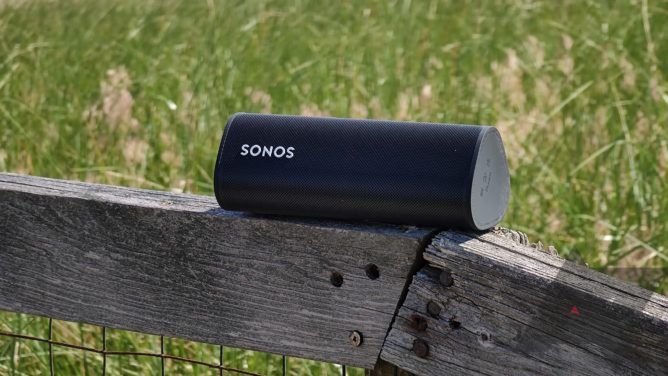 Um alto-falante Sonos Roam colocado em uma cerca de madeira com grama ao fundo