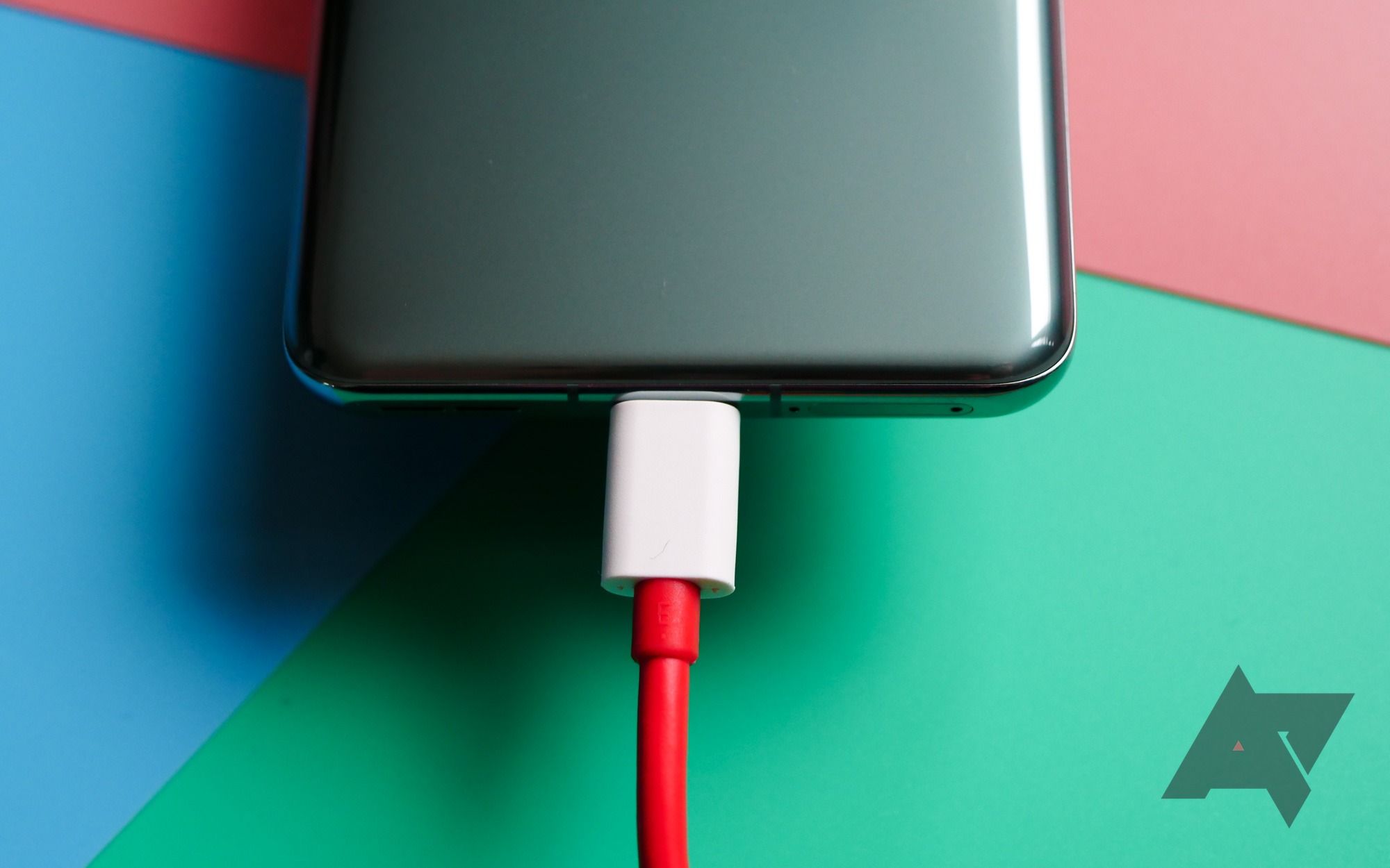 O terço inferior traseiro de um smartphone OnePlus 11 paira sobre um fundo de papelão tricolor com um cabo USB-C branco e vermelho conectado