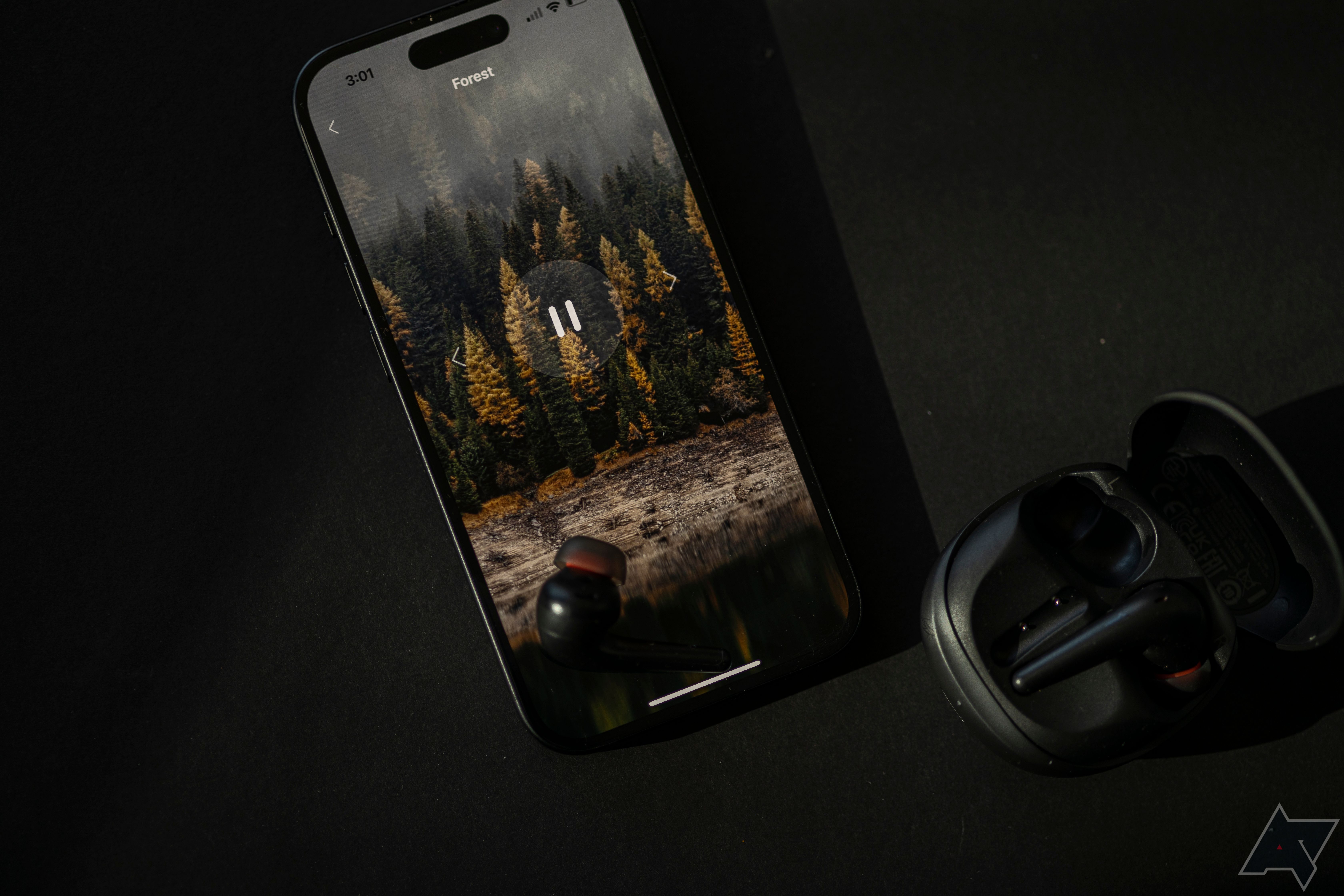 1Mais fone de ouvido Aero colocado no iPhone, aberto para a paisagem sonora da floresta, com capa de fone de ouvido aberta por perto