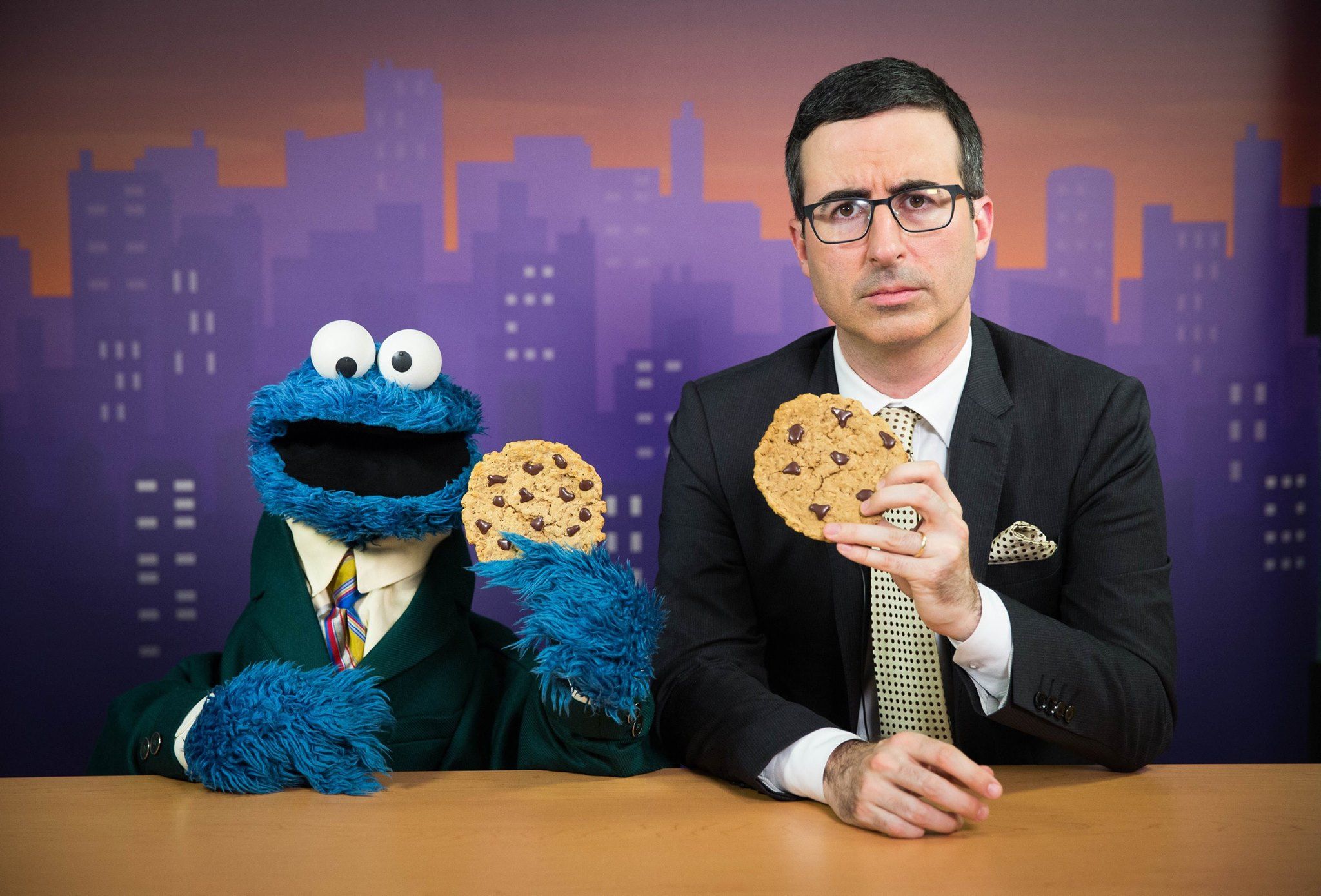 O Cookie Monster e John Oliver sentados em uma mesa segurando biscoitos