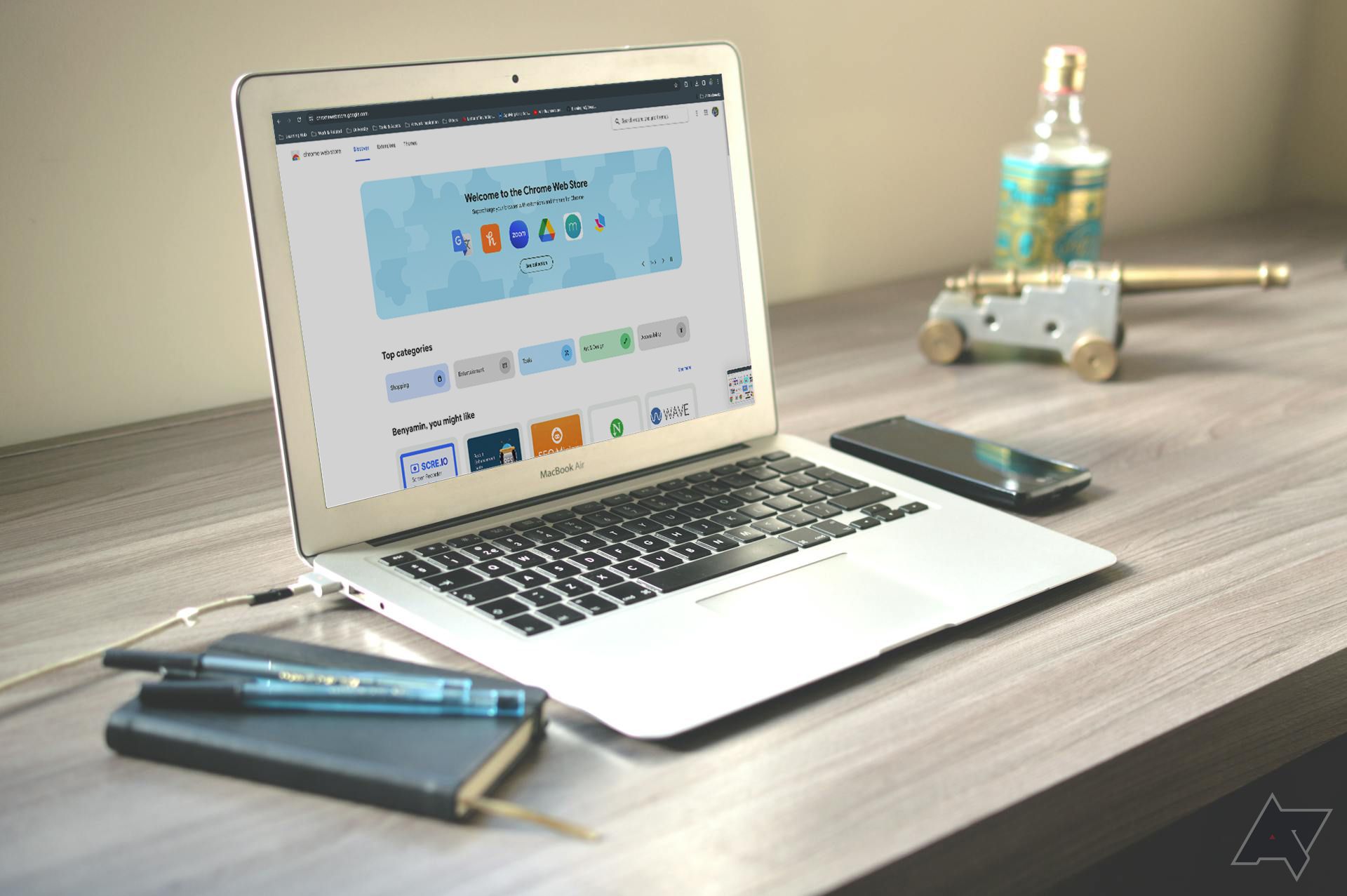 Um laptop em uma mesa de madeira exibindo a página inicial da Chrome Web Store, com um smartphone, uma garrafa, canetas e um canhão em miniatura ao lado.