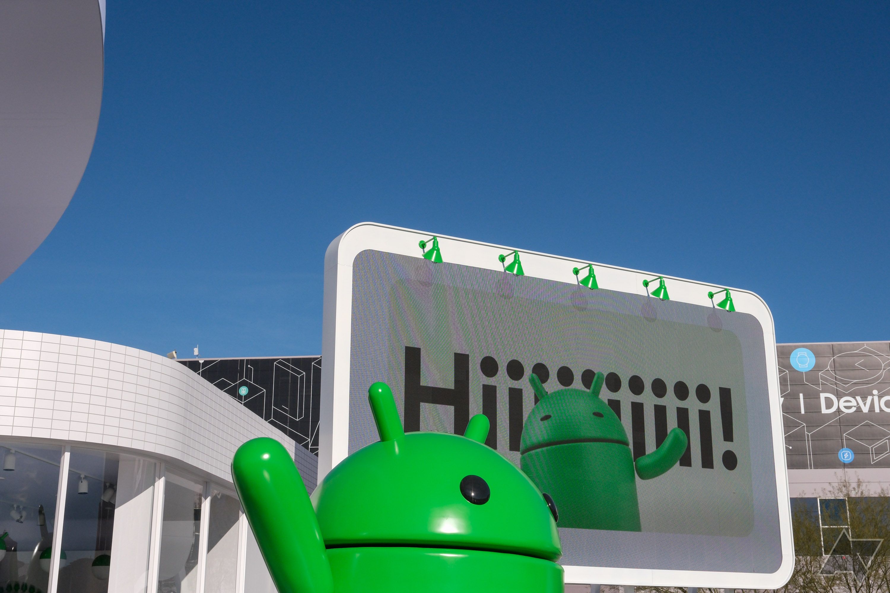 Um mascote Android em frente a um outdoor onde se lê 'Hiiiiiiiiii!'