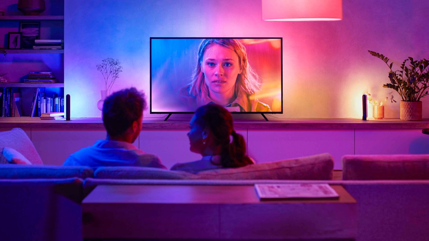 Um casal sentado em um sofá de costas para a câmera, assistindo TV com iluminação ambiente combinando com o conteúdo da tela