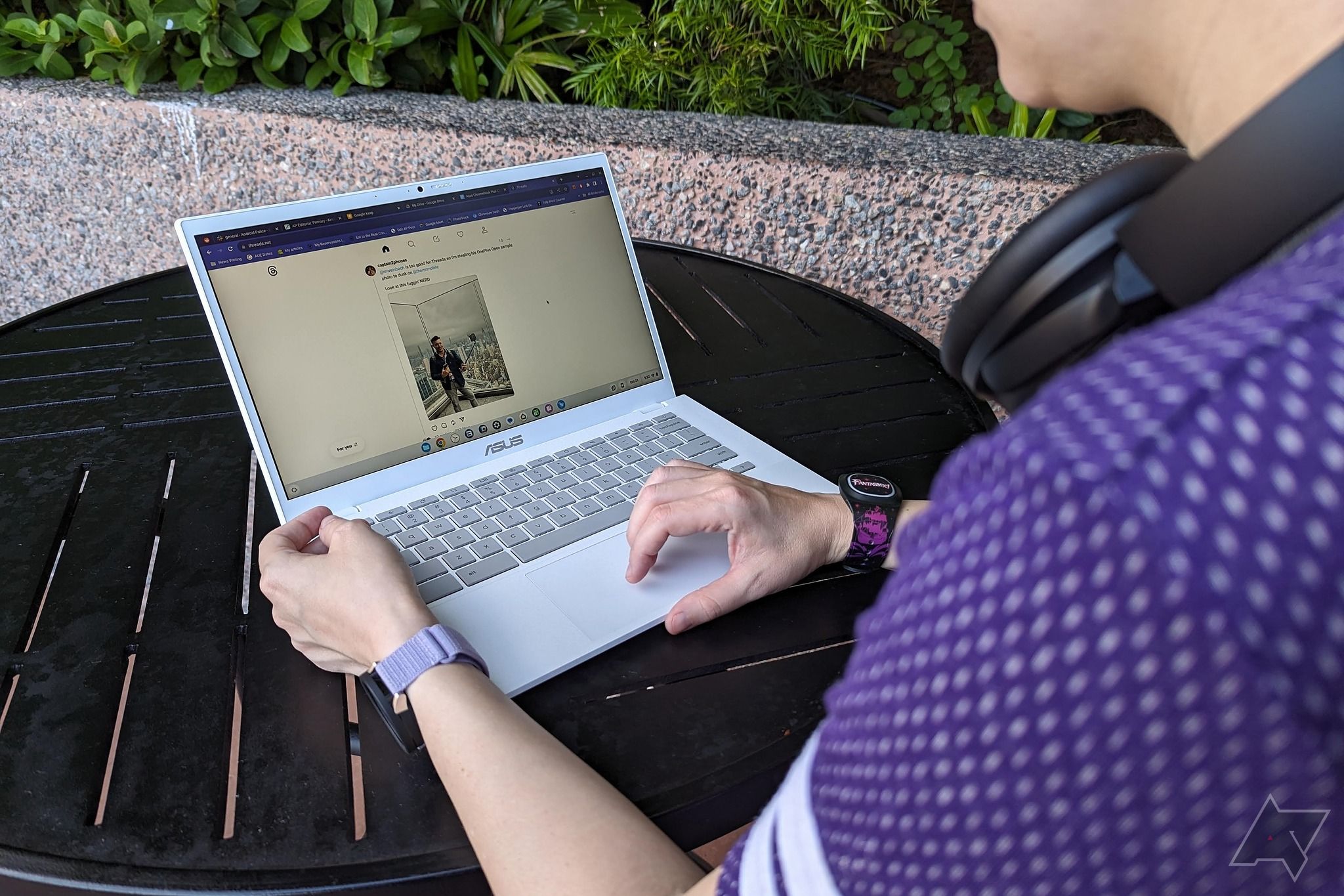 Um Asus Chromebook Plus CX34 em uma mesa preta do lado de fora sendo usado por uma pessoa com uma camisa roxa e fones de ouvido