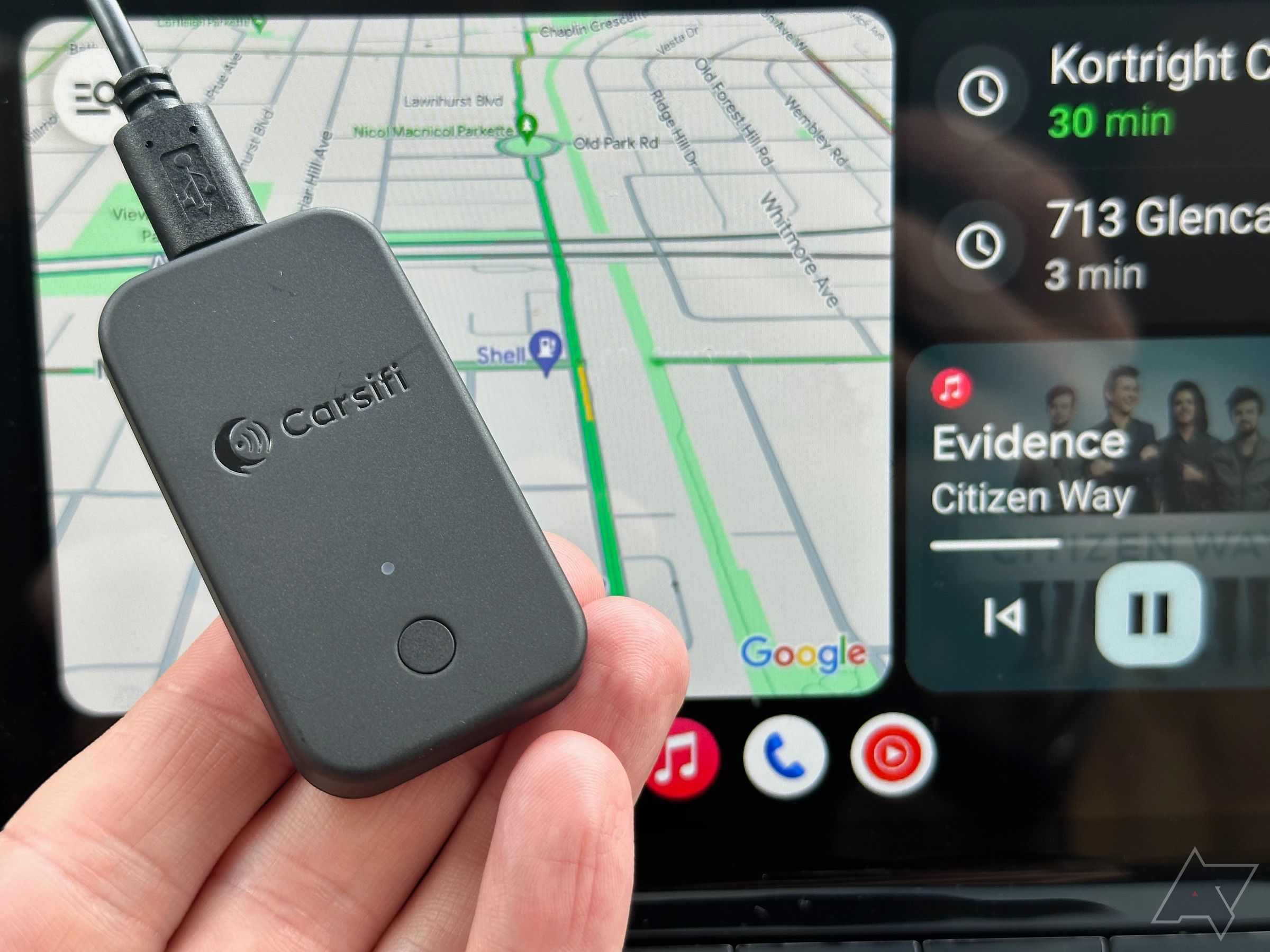 Dedos segurando o adaptador Carsifi Wireless Android Auto na frente da tela do Android Auto mostrando o Google Maps e o Apple Music.