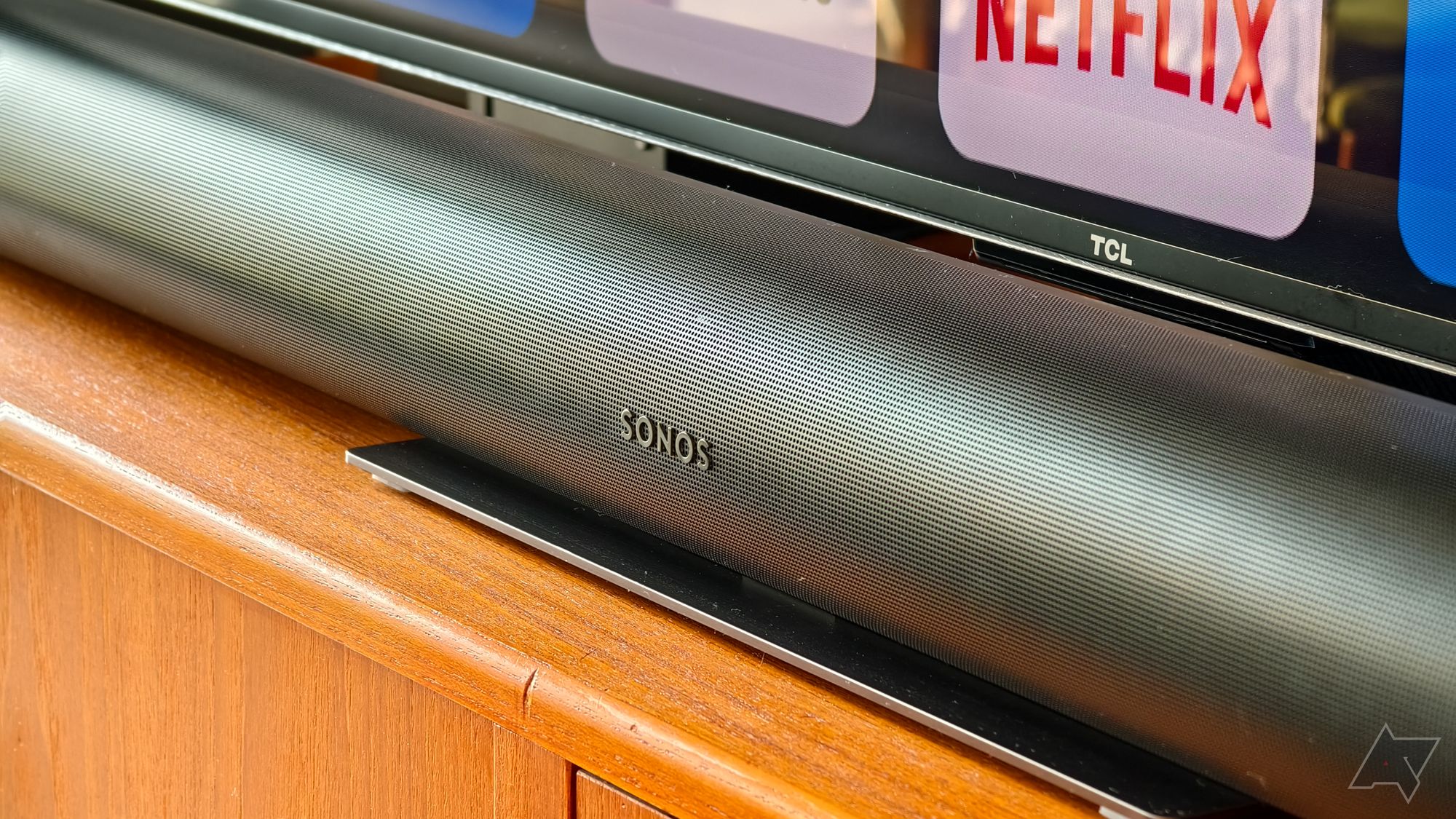 Barra de som Sonos Arc em preto, fotografada acima e de um ângulo, sentada em um suporte de TV de teca com a parte inferior da TV TCL visível atrás mostrando um logotipo da Netflix