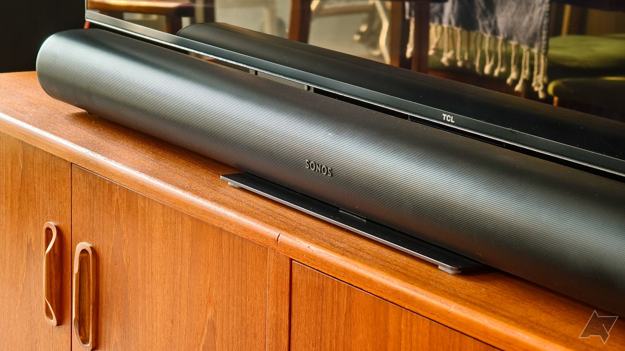 Uma foto de uma barra de som Sonos Arc preta em um suporte de TV de madeira teca, abaixo de uma TV TCL, com as borlas de um cobertor visíveis refletidas na TV