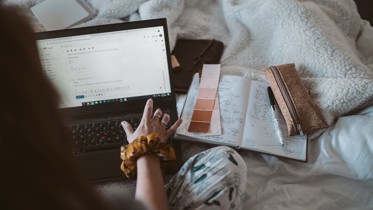 Pessoa sentada na cama com o laptop aberto, com algumas amostras de tinta e um caderno à esquerda. 