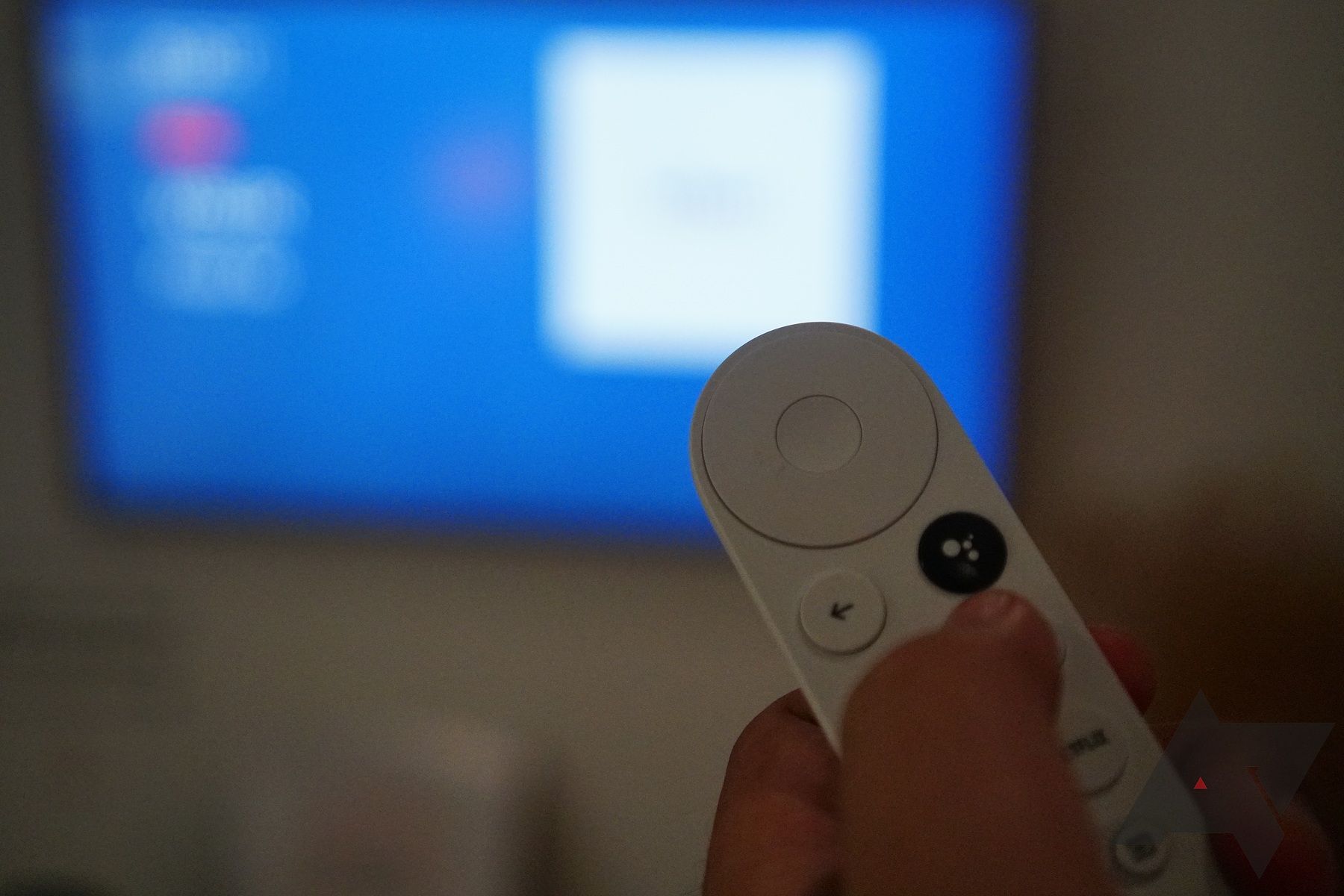 Mão usando um controle remoto branco do Google Chromecast com uma TV desfocada ao fundo.