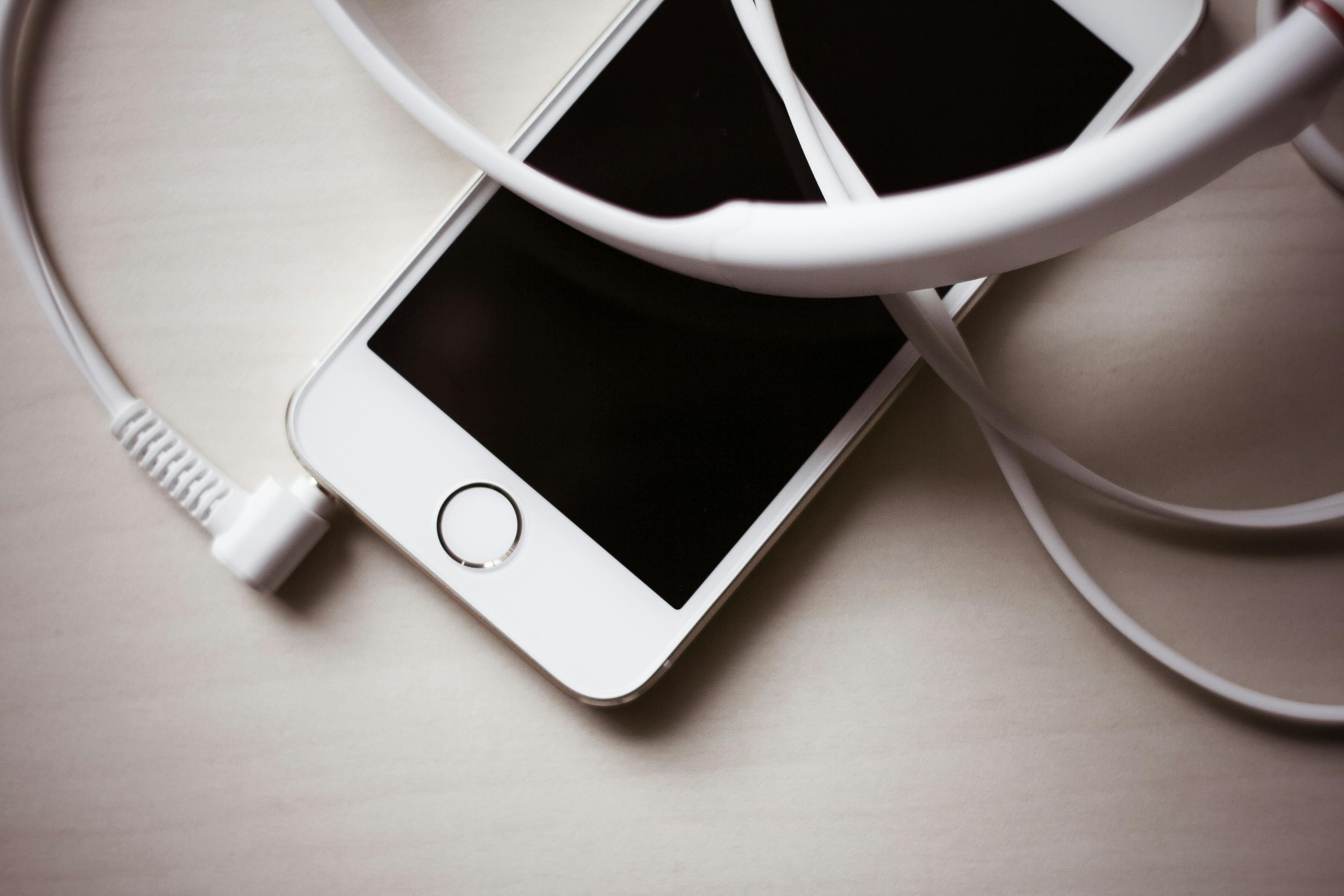 O iPhone 5s com botão home está em uma superfície, emaranhado em seu cabo de carregamento branco, exibindo uma tela preta indicando que está desligado ou em modo de espera.