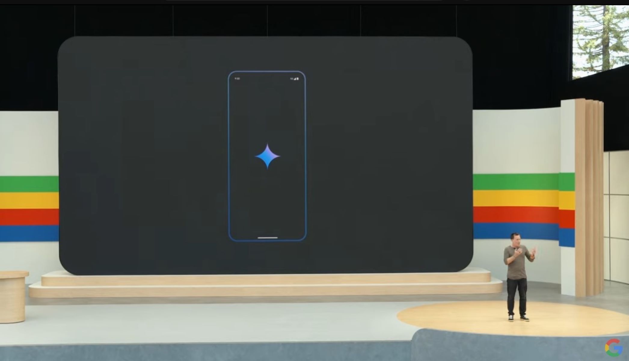 Captura de tela de um homem no palco do Google I/O com uma imagem do aplicativo Google Gemini em um Pixel atrás dele.