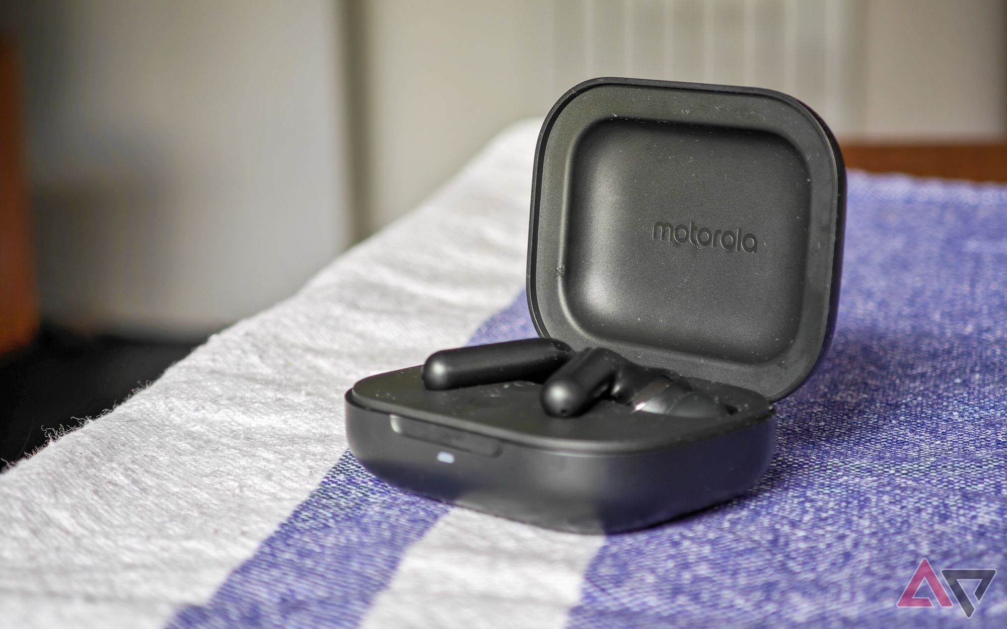 Os Moto Buds+ em preto com os fones de ouvido no estojo de carregamento aberto sobre uma toalha de mesa azul e branca