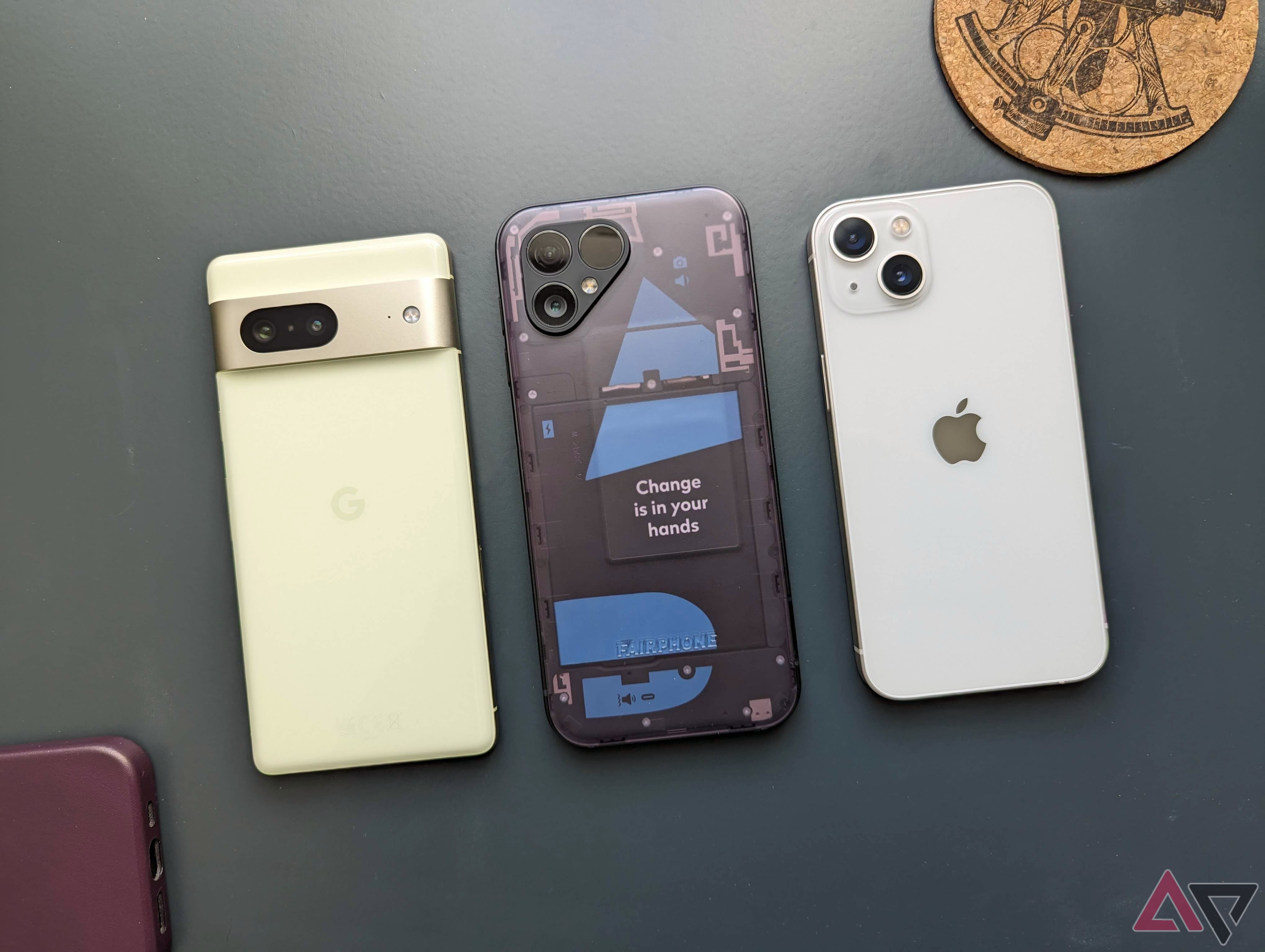O Google Pixel 7, o Fairphone 5 e o Apple iPhone 13 próximos um do outro em um fundo verde com itens aleatórios espalhados ao redor deles