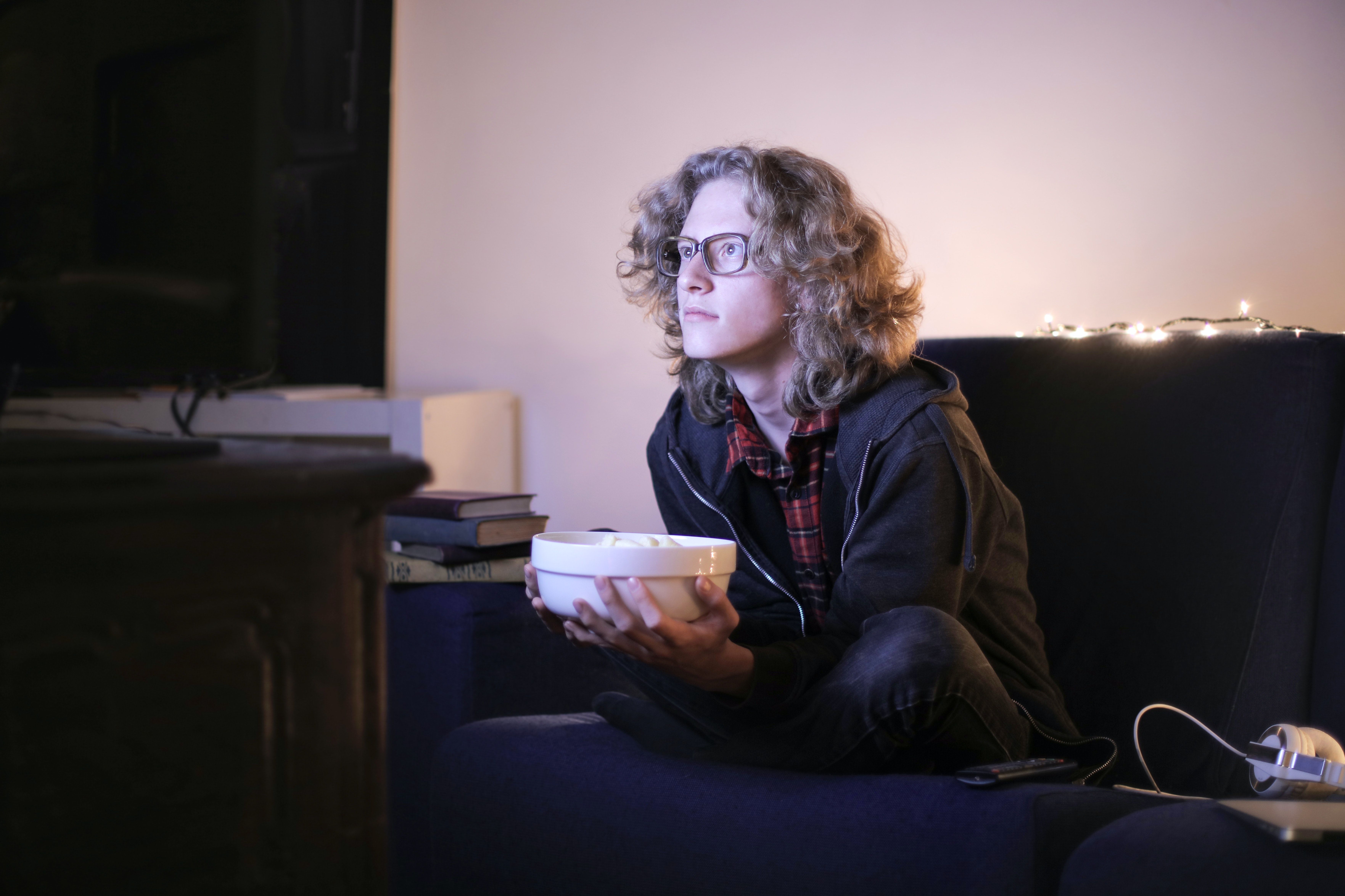 Um homem de cabelos compridos e óculos segura uma tigela de pipoca enquanto assiste televisão. 