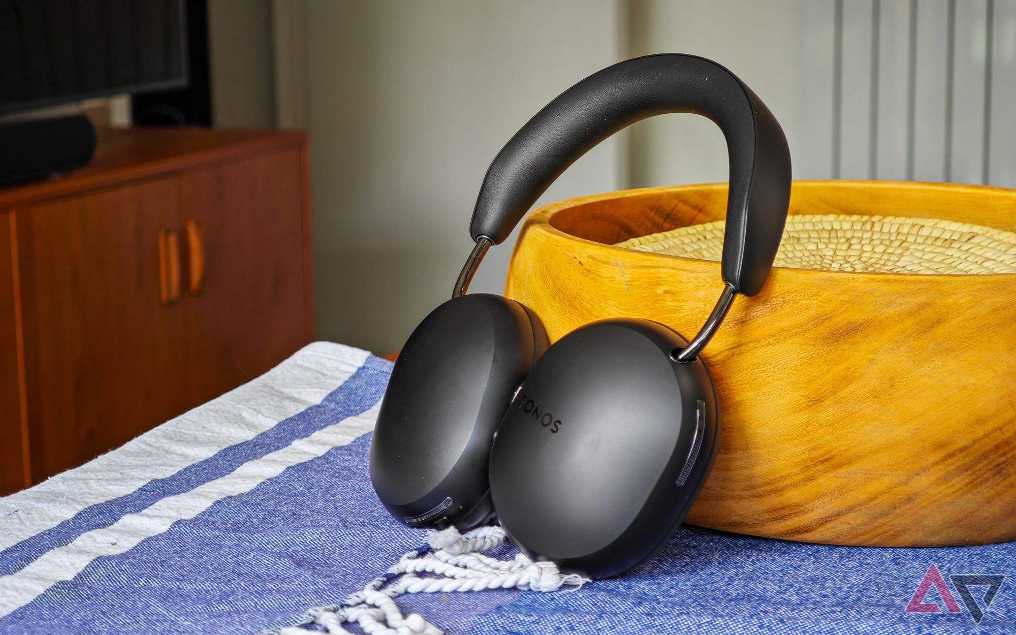 Fones de ouvido Sonos Ace apoiados em uma tigela de madeira sobre uma mesa com uma toalha de mesa azul