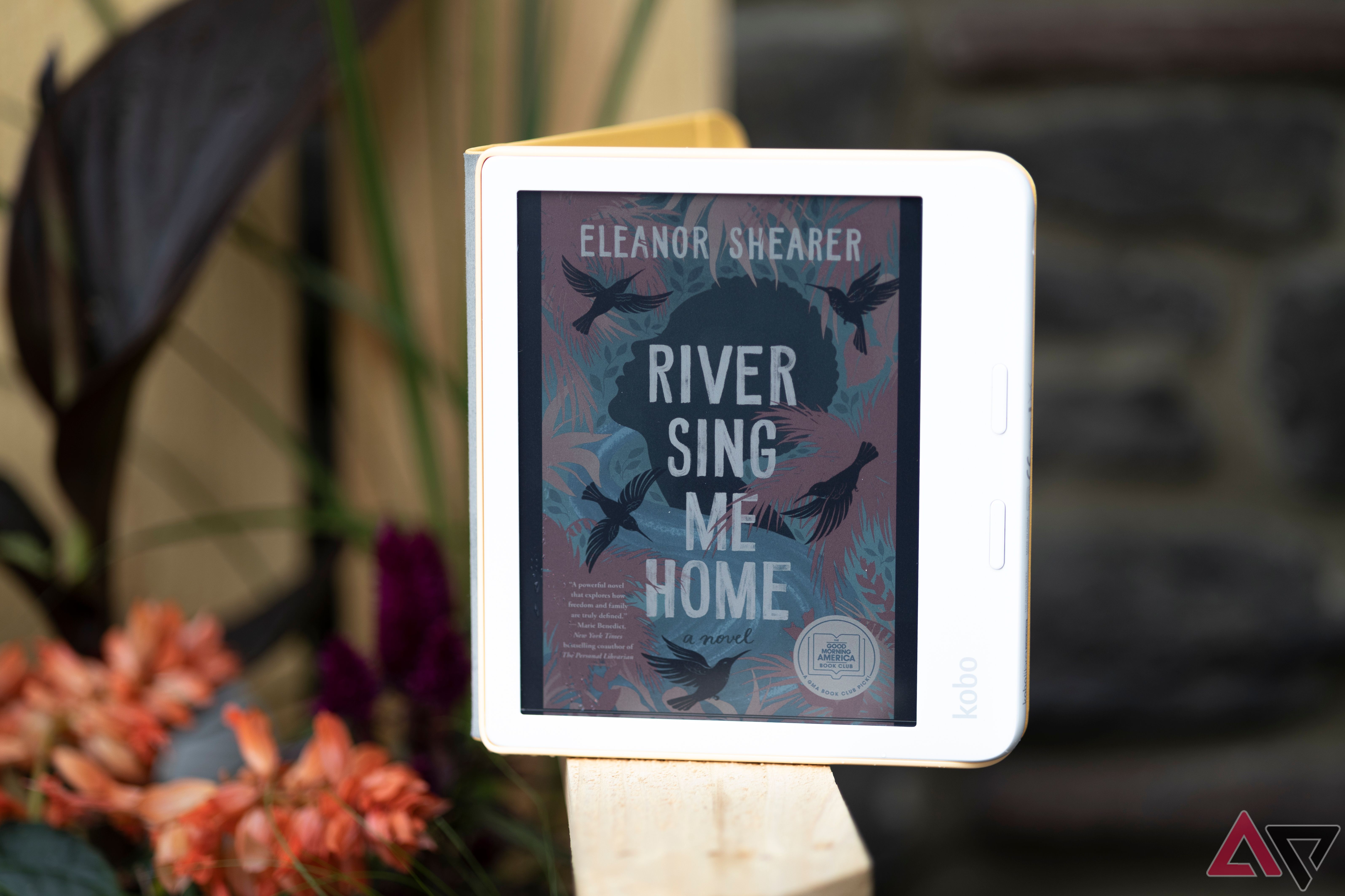 Kobo Libra Color abre o cover de 'River Sing Me Home' apoiado em um banco de madeira