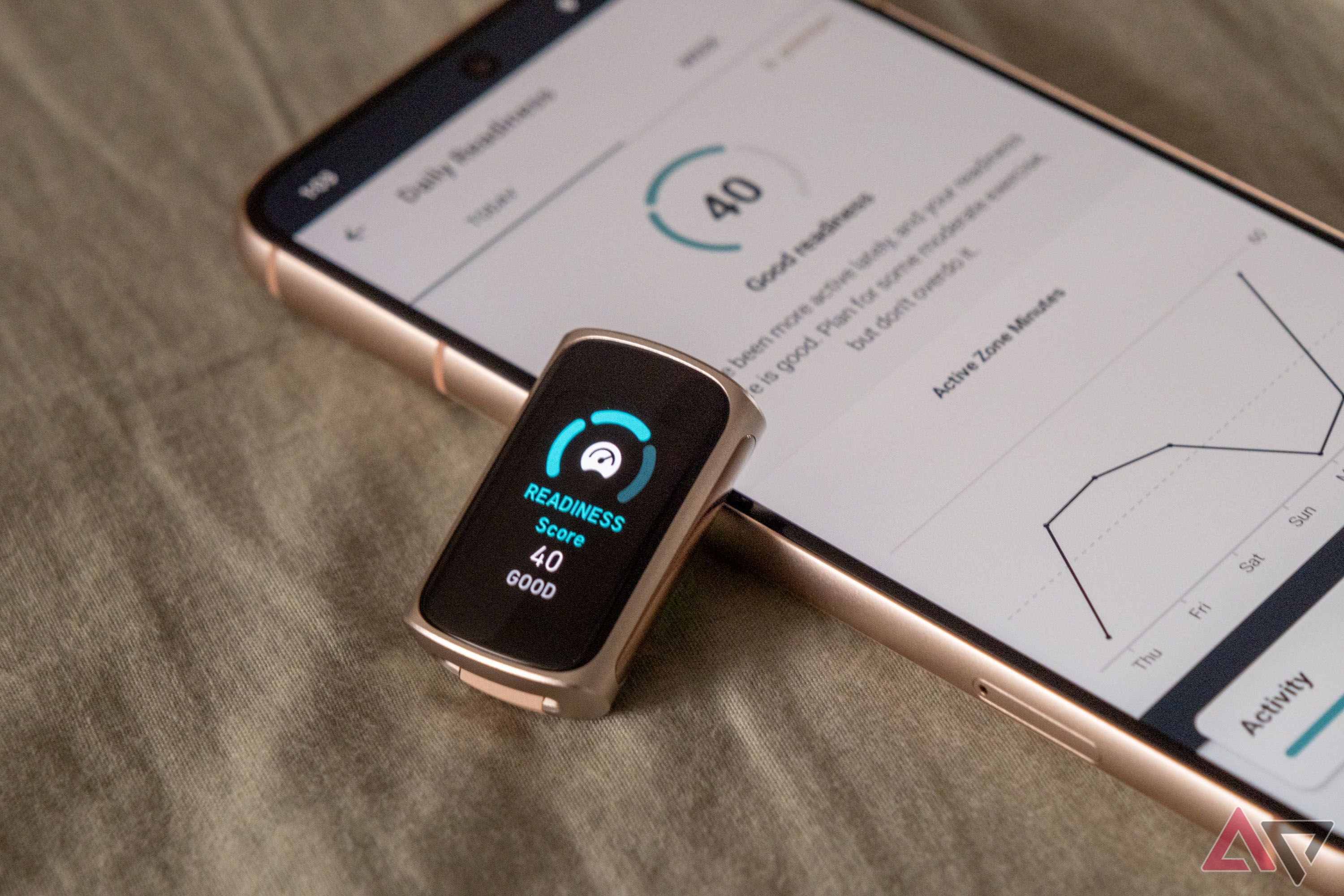 Um Fitbit Charge 6 sem faixas mostrando uma pontuação de prontidão apoiado em um smartphone com o aplicativo Fitbit aberto na página de pontuação de prontidão em uma mesa de madeira.