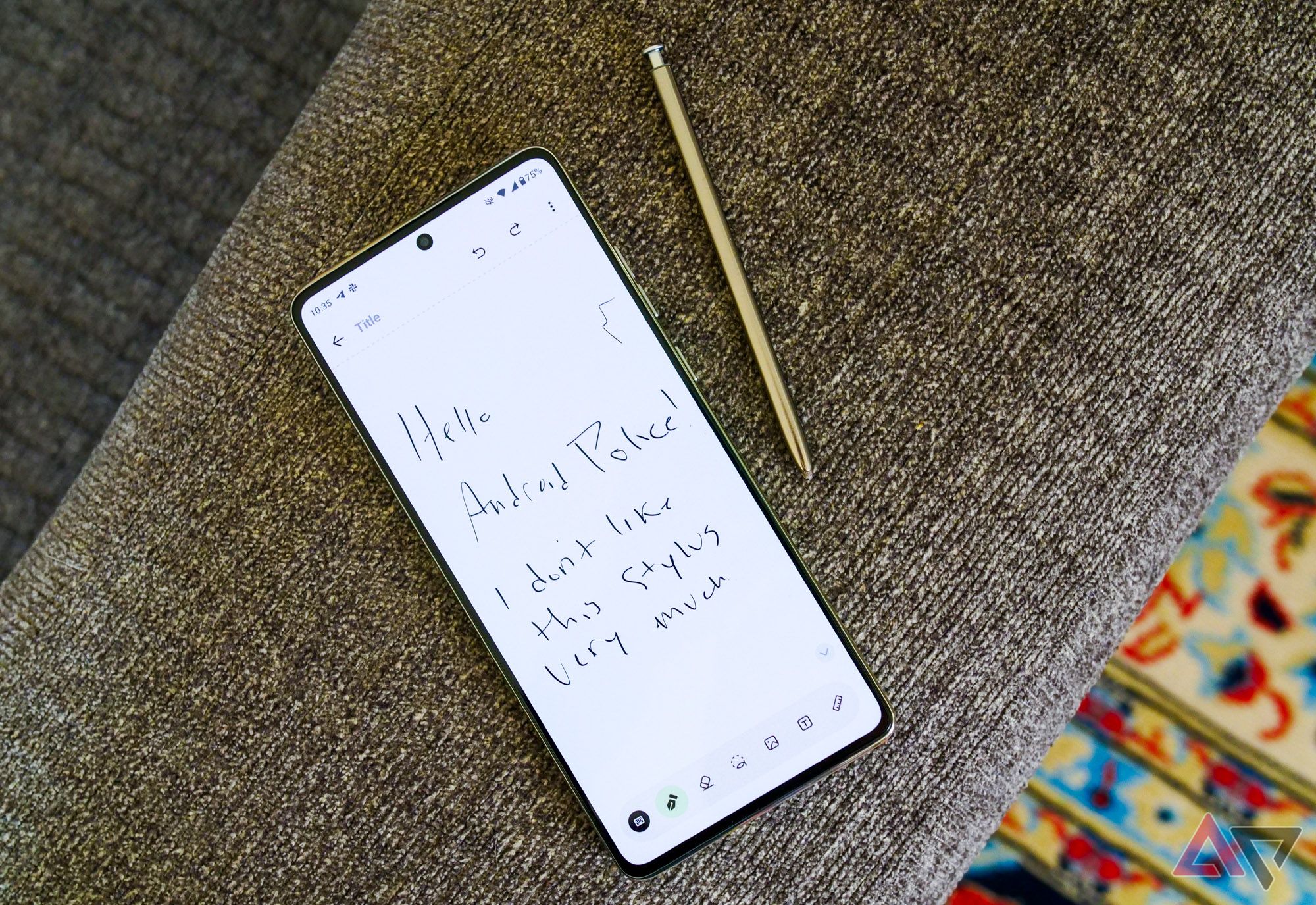 O Moto G Stylus 2024 sentado no braço do sofá com a caneta para fora e "Olá Android Police! Não gosto muito desta caneta" escrito na tela.