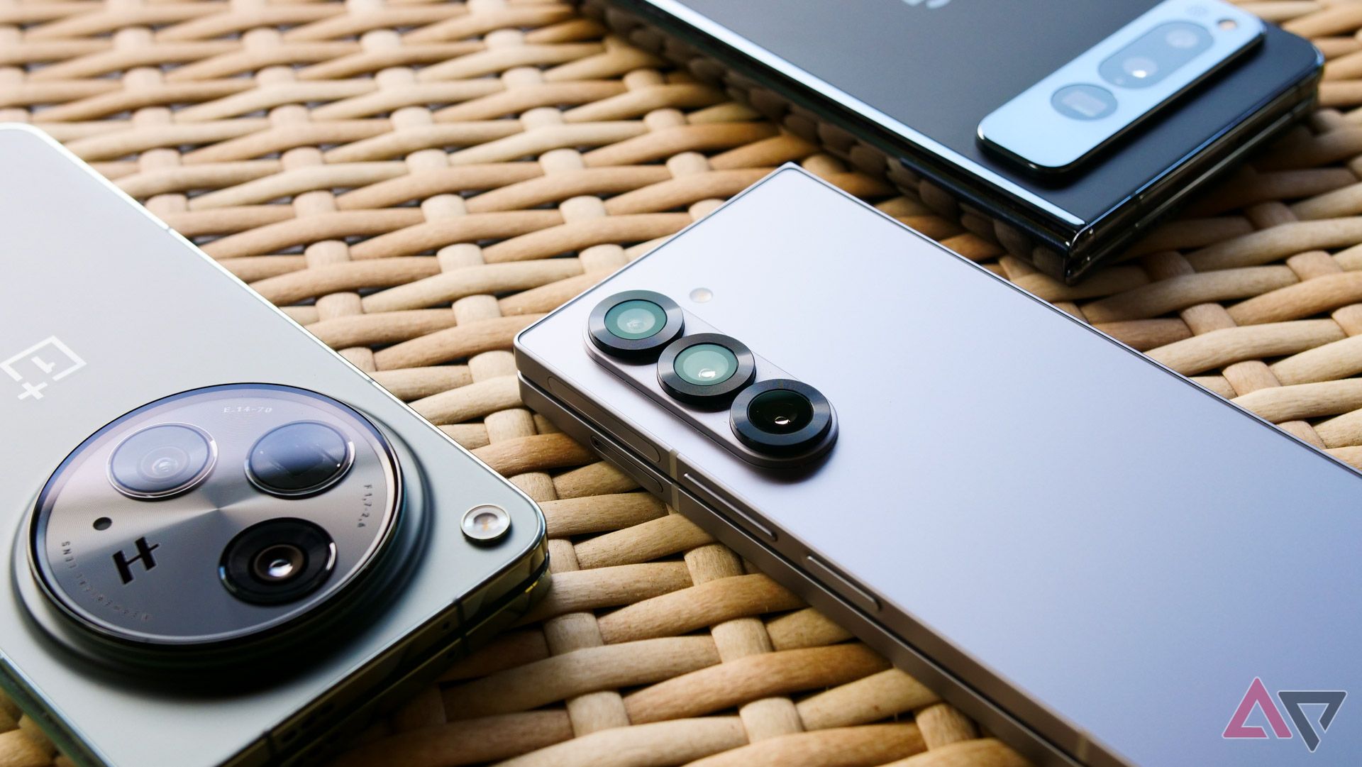 O Galaxy Z Fold 6, o OnePlus Open e o Pixel Fold sobre uma mesa de vime com suas saliências de câmera mostradas.