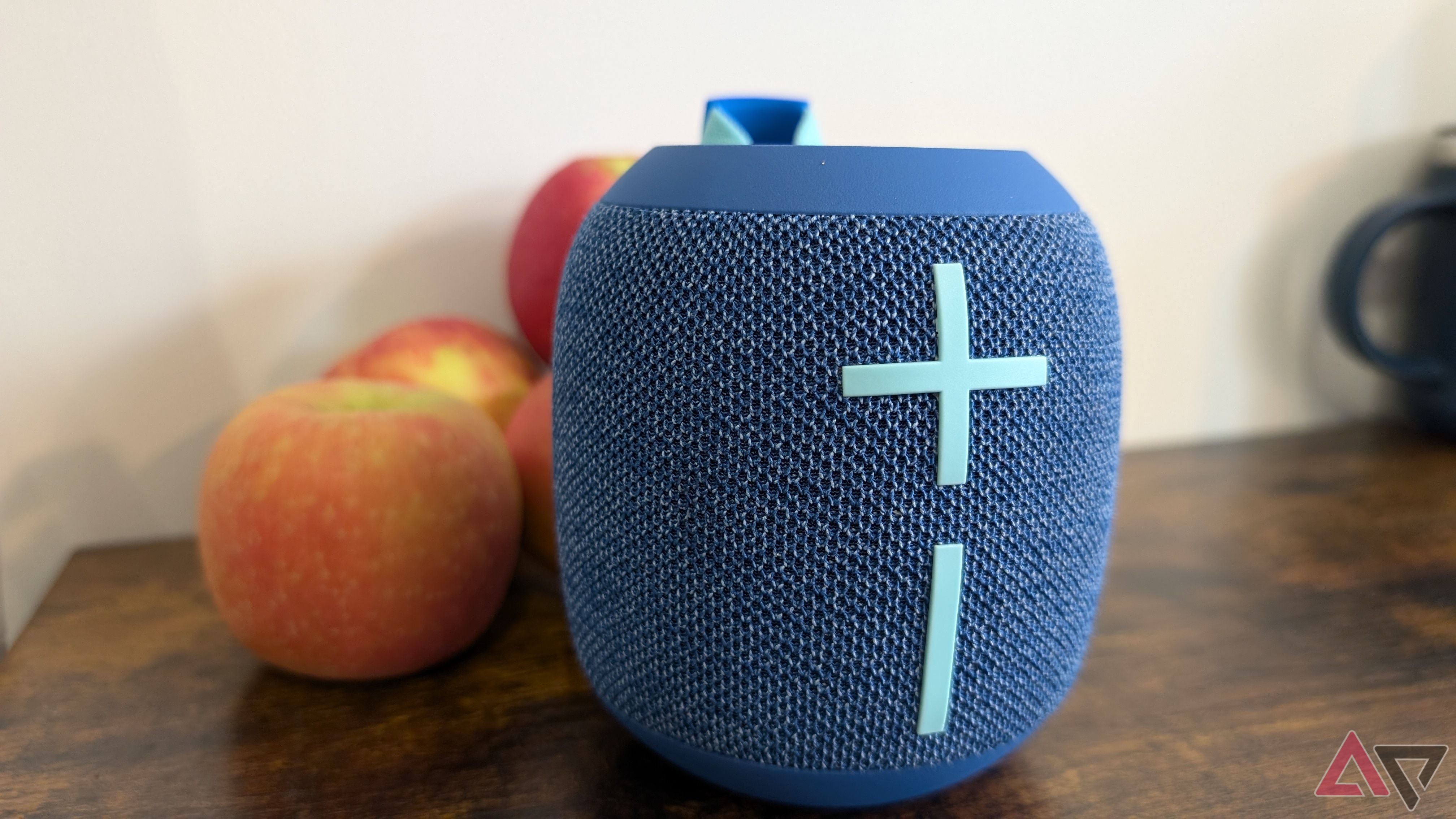 alto-falante bluetooth azul com botões mais e menos azul claro em mesa de madeira em frente a maçãs