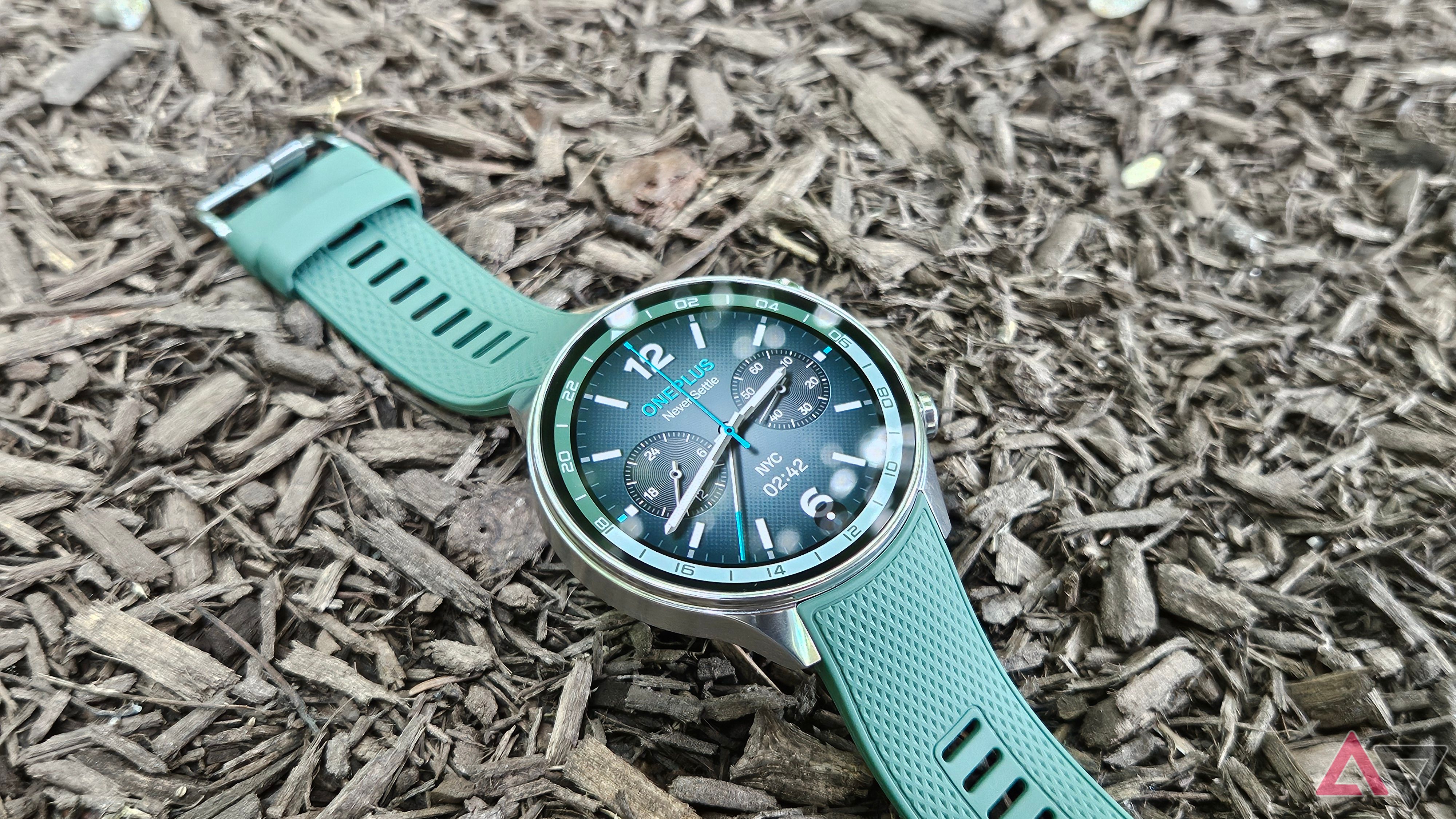 OnePlus Watch 2R em Forest Green apoiado em Mulch com um relógio analógico