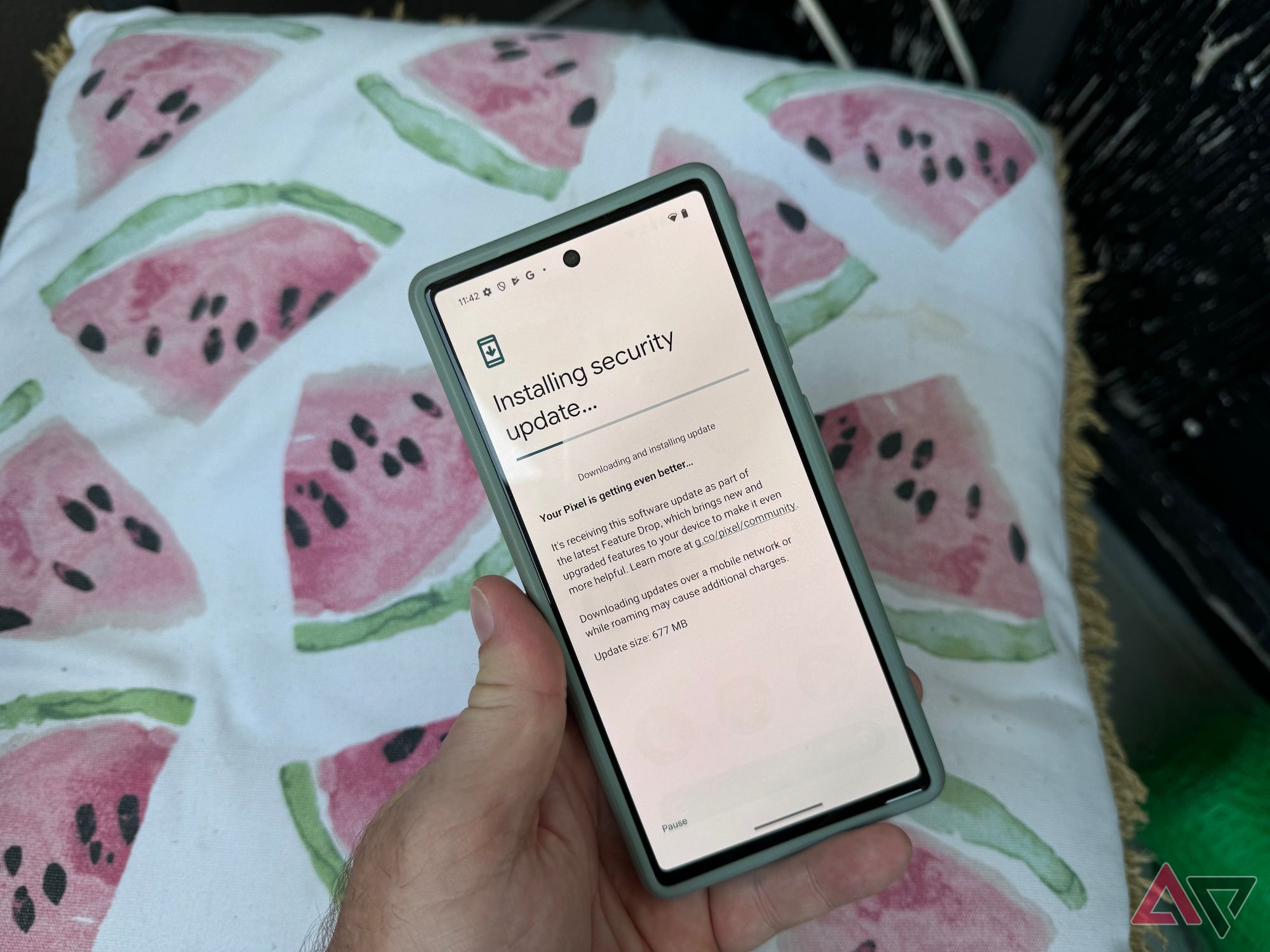 Mão de um homem segurando um Pixel 6 mostrando uma atualização em andamento com um travesseiro ao fundo com imagens de fatias de melancia rosa.