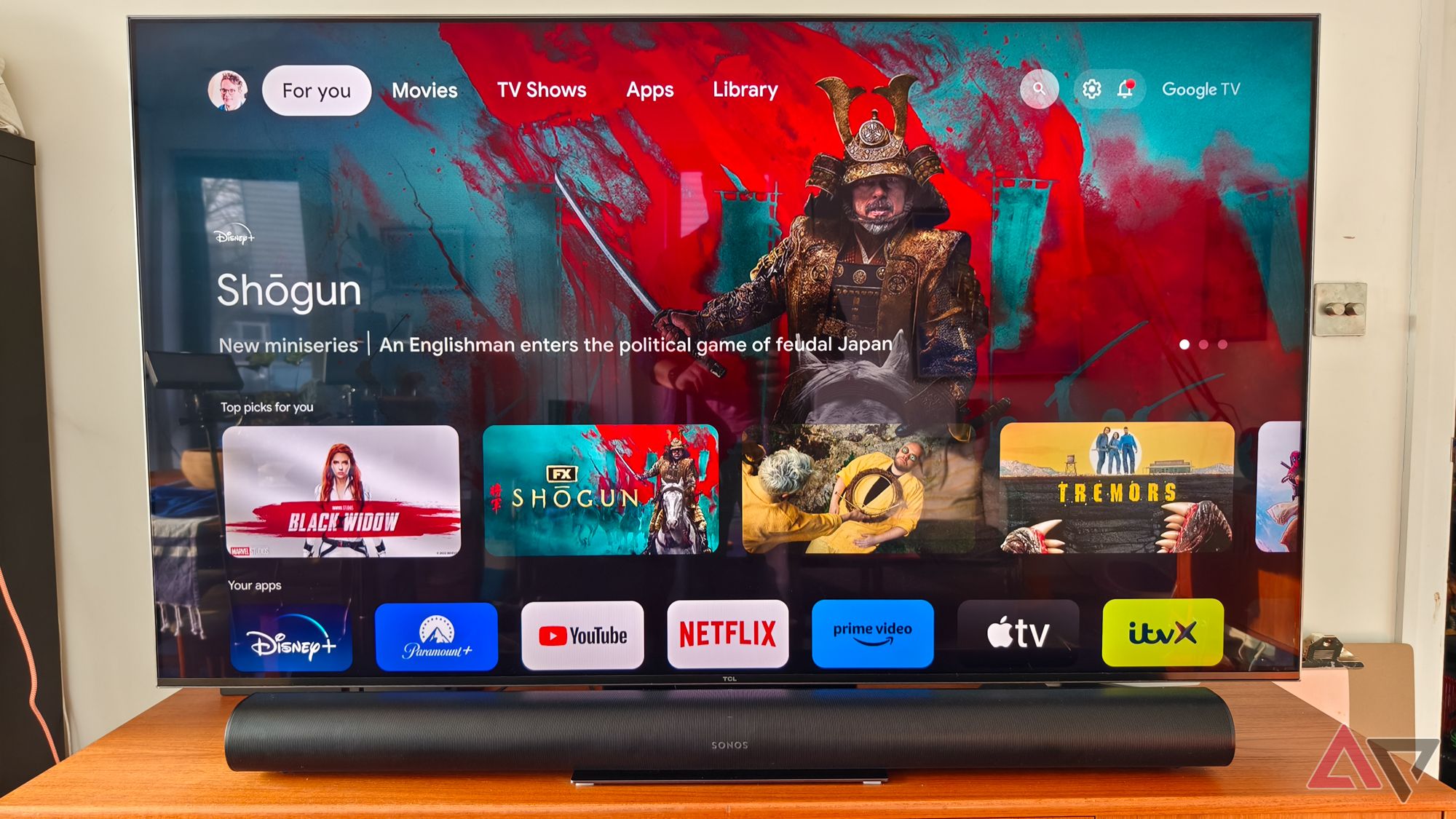 Uma foto frontal de uma barra de som Sonos Arc preta em um suporte de TV de teca abaixo de uma TV TCL. A TV está mostrando a tela inicial do Google TV com um grande anúncio da série de TV Shogun