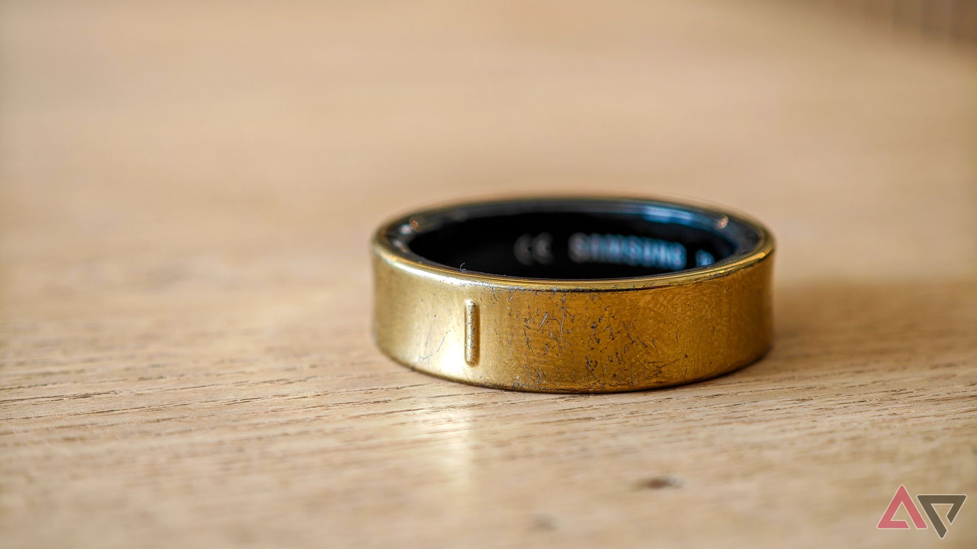 Um Samsung Galaxy Ring dourado deitado de lado sobre uma mesa de madeira clara com alguns arranhões visíveis no acabamento