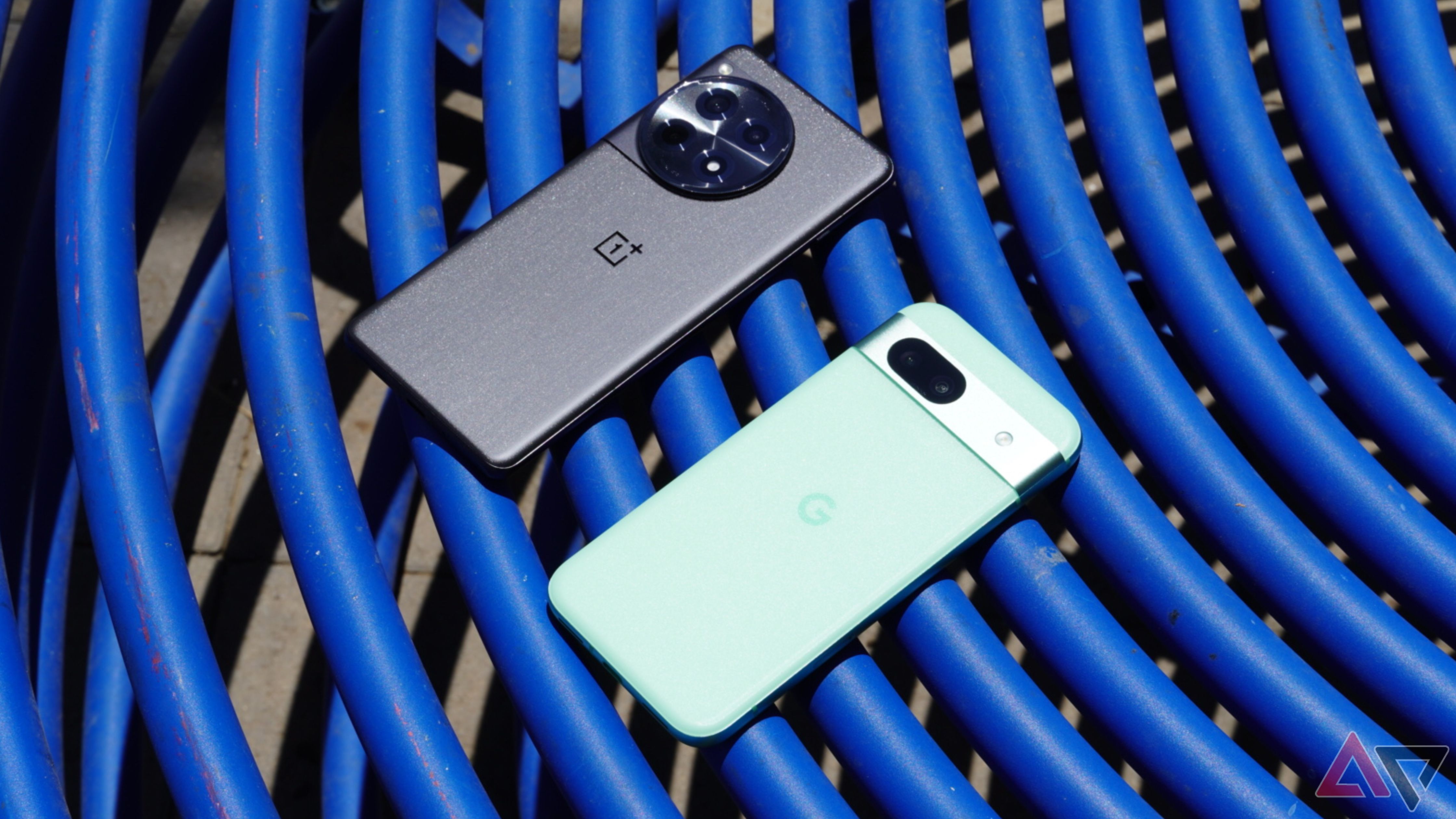 O OnePlus 12R e o Pixel 8a em uma mesa azul ao ar livre.