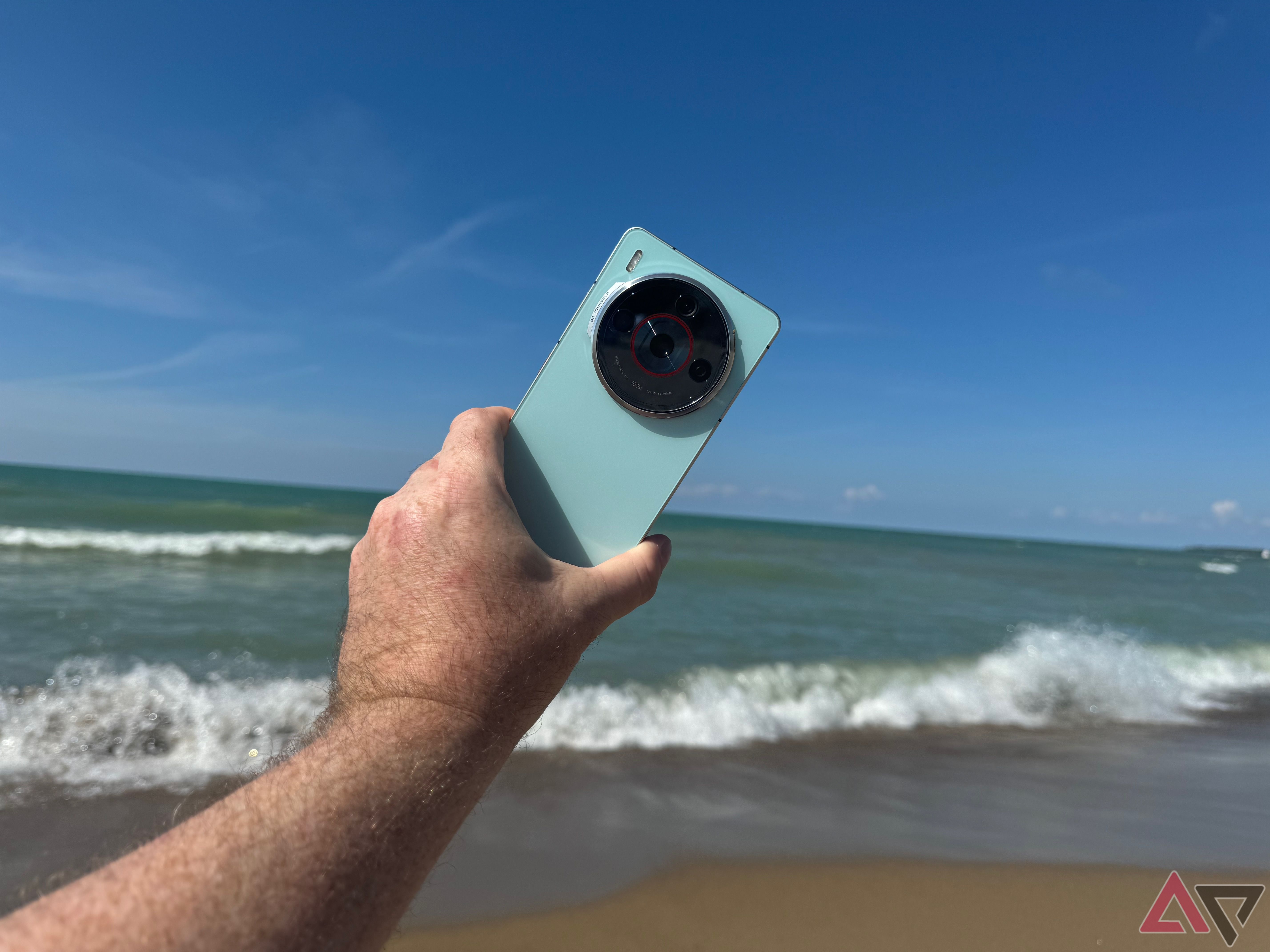 Mão de um homem segurando uma Nubia Z60S Pro azul-turquesa no ar em uma praia com ondas ao fundo.