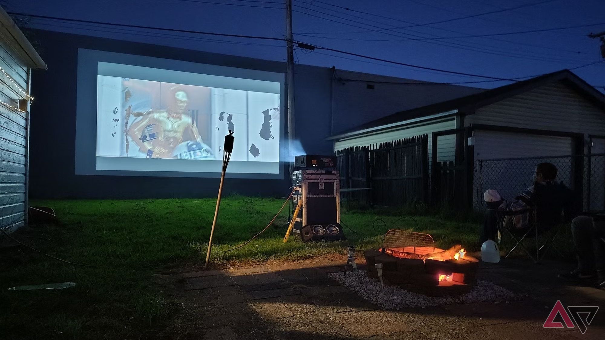 Uma cena ao ar livre mostrando uma fogueira e um filme passando em um projetor.