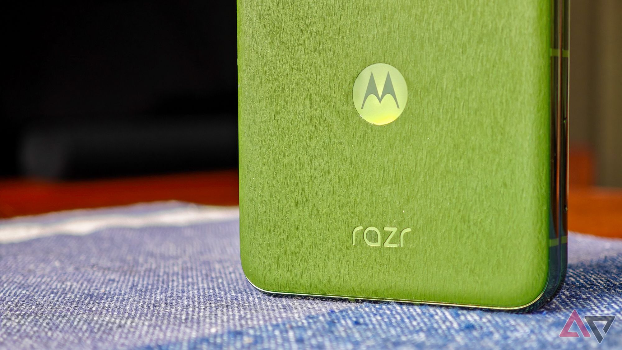 Motorola Razr+ 2024 apoiado sobre uma toalha de mesa azul, fechado, mostrando a traseira verde com close do logotipo Razr