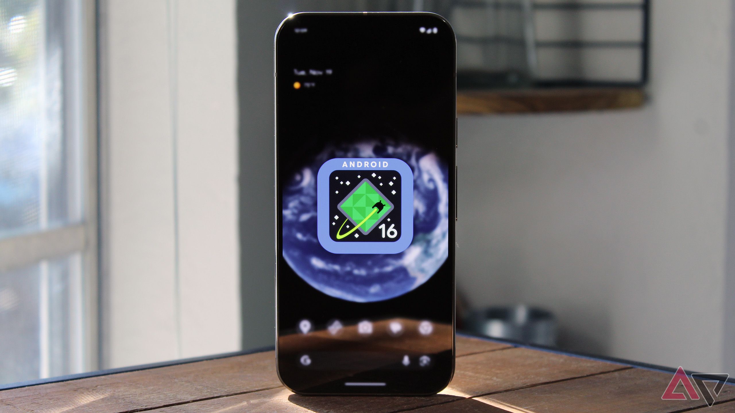 Imagem do logotipo do Android 16 na tela inicial de um Pixel 9 Pro com o telefone em cima de uma mesa de madeira.