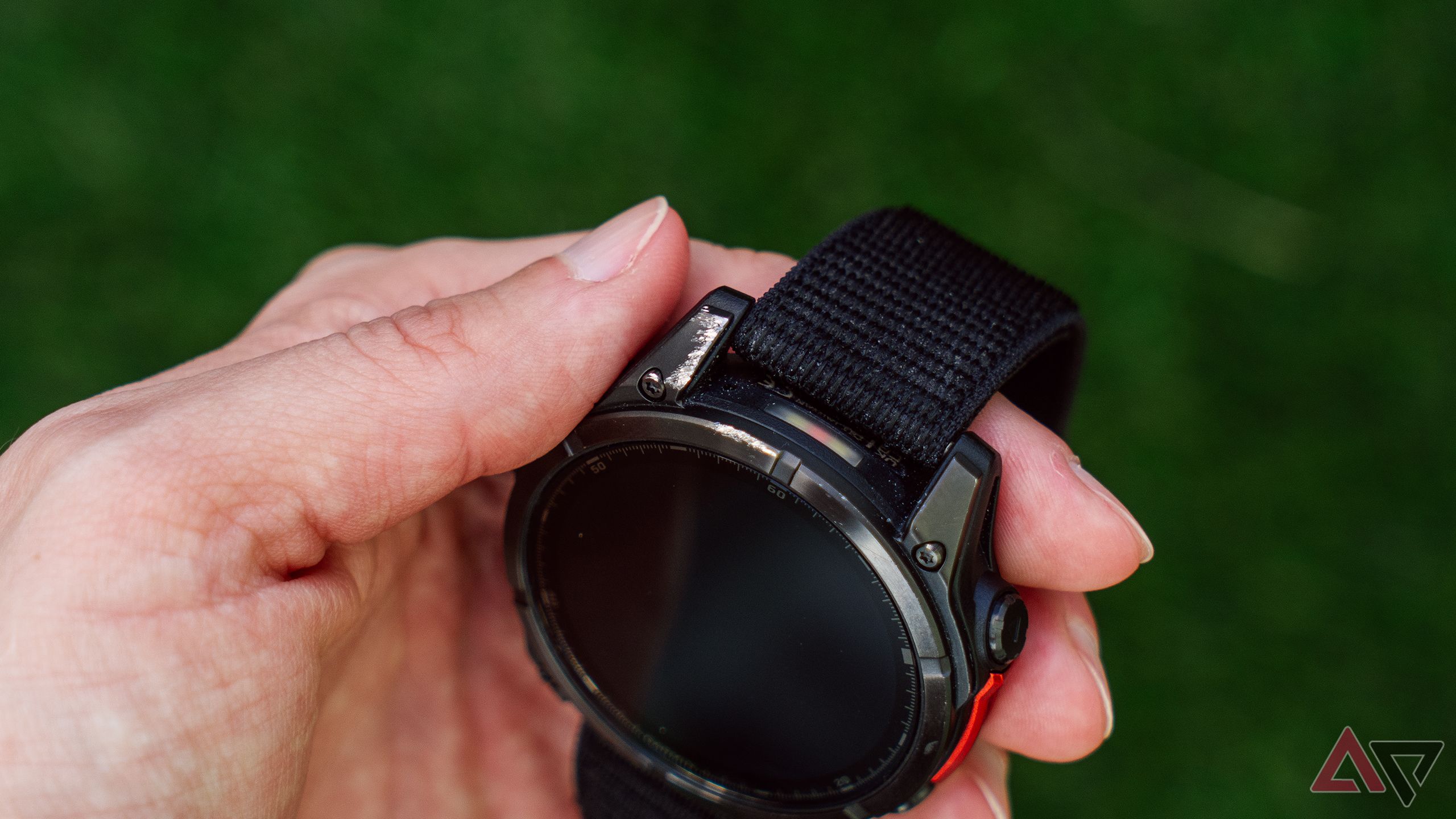 Uma mão segura um Garmin Fenix ​​8 AMOLED arranhado acima de um fundo verde desfocado. 