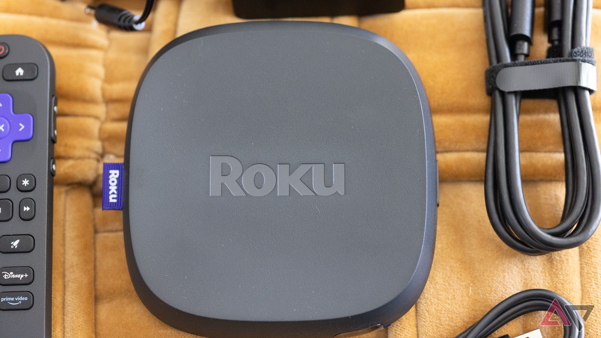 A caixa de streaming Roku Ultra 2024 em um fundo amarelo ao lado de um controle remoto e cabos