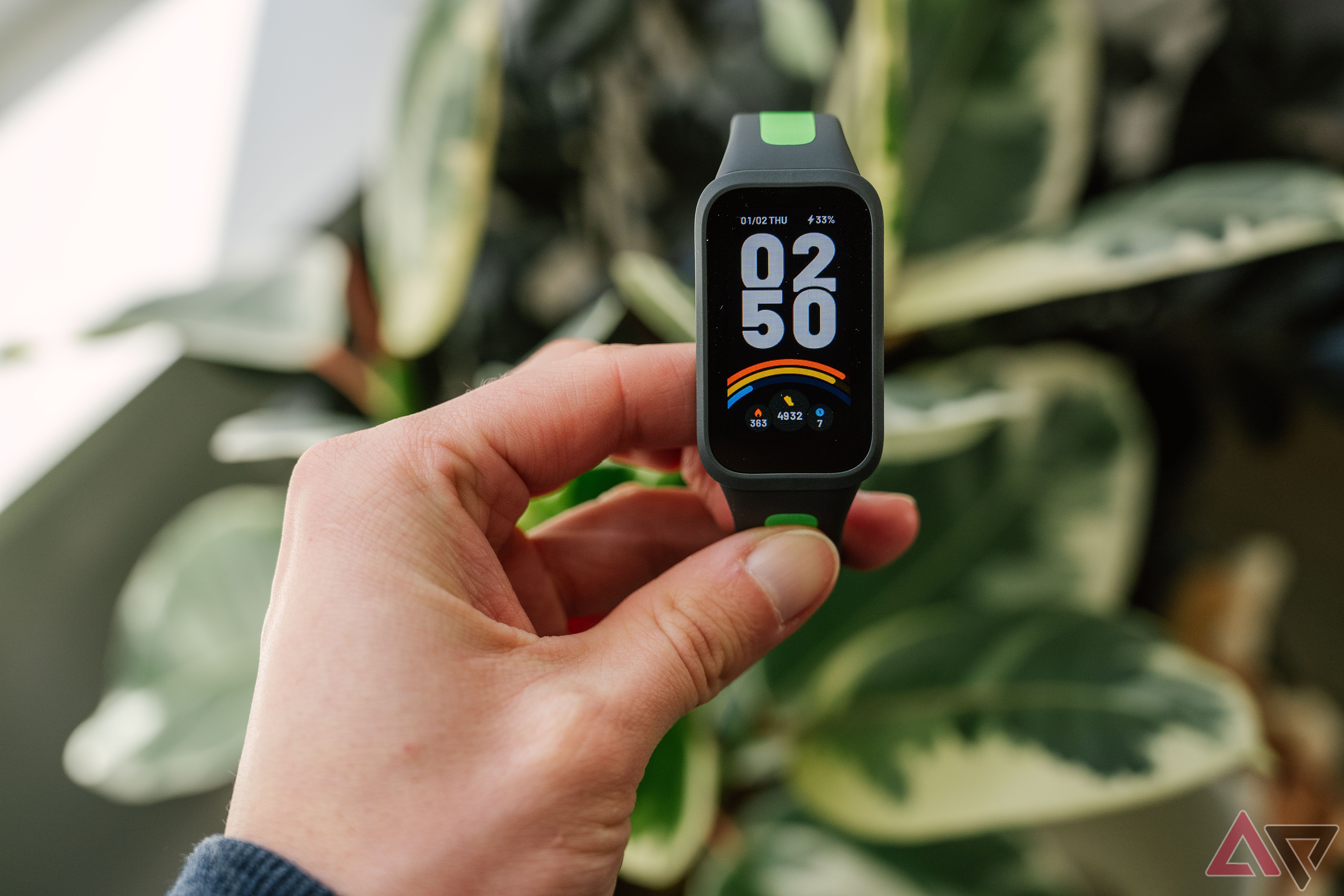 Uma mão segura a Xiaomi Smart Band 9 Active acima de plantas verdes desfocadas. 